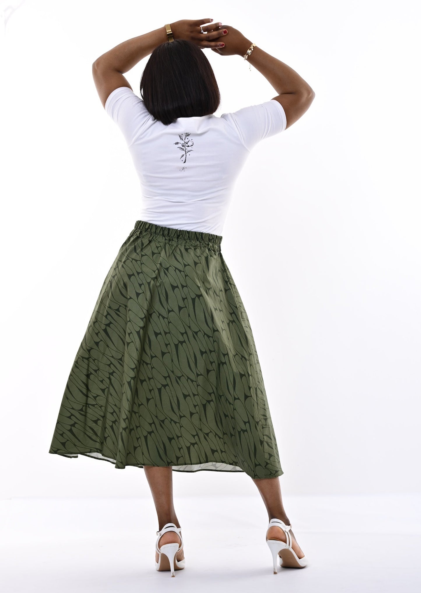A woman is shown from behind wearing a white short-sleeve t-shirt and a knee-length olive green skirt with a subtle pattern, standing against a bright white studio background.
