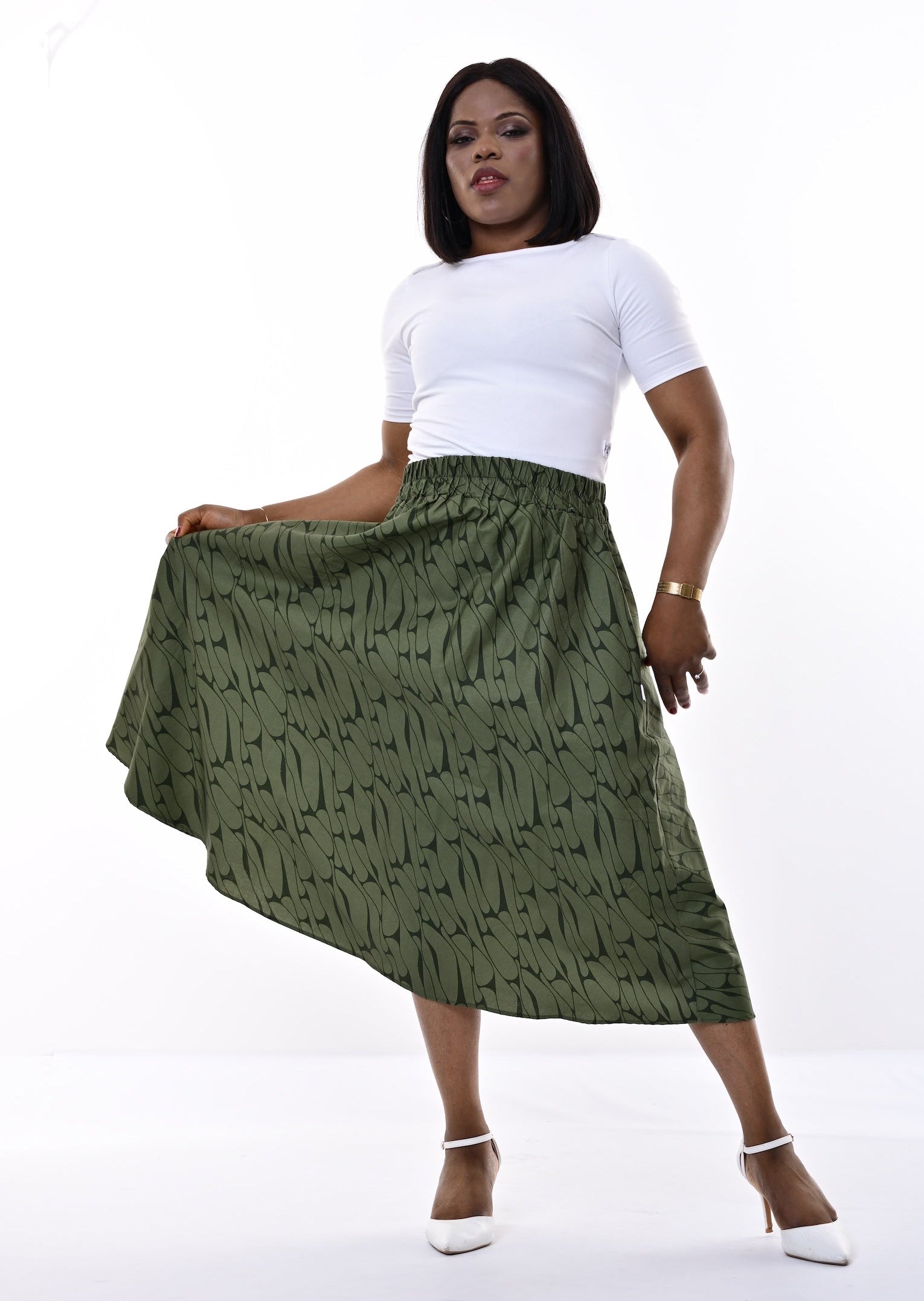 A woman modeling a high-waisted olive green midi skirt with a black abstract brushstroke pattern, paired with a white short-sleeve top and white heels, against a white studio background.