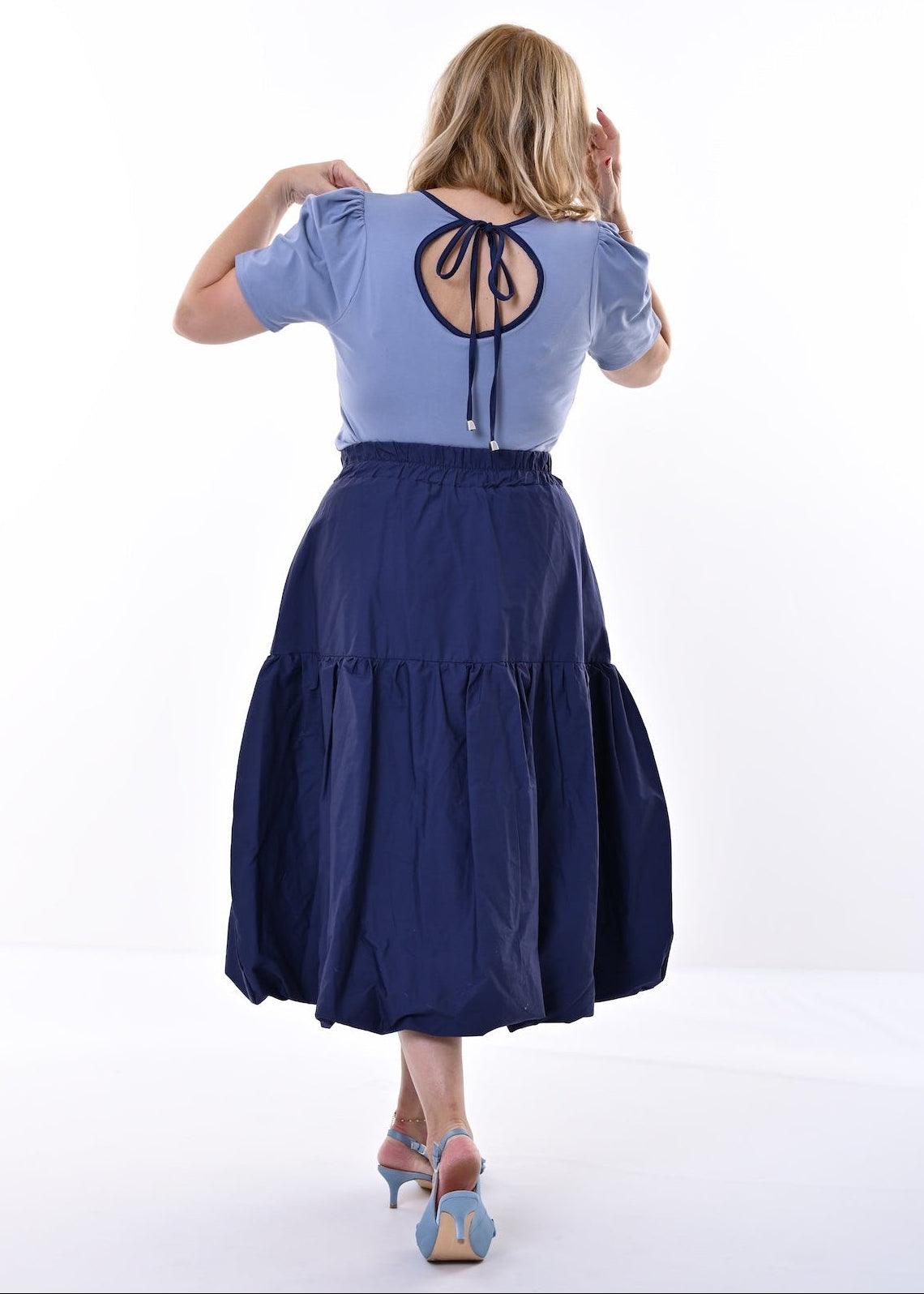 A woman shown from the back wearing a light blue short-sleeve top with a circular lace-up back detail and a dark navy midi skirt, standing against a plain white studio background.