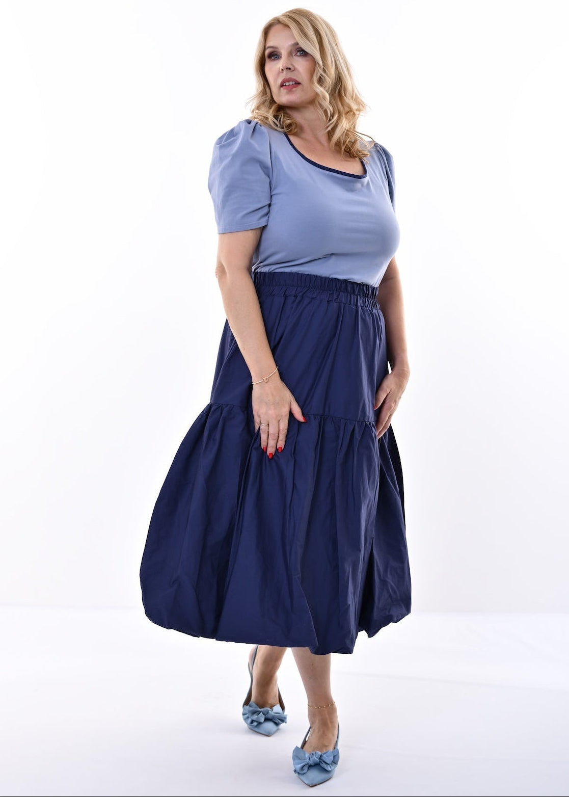 A woman modeling a two-piece outfit consisting of a light blue short-sleeve top and a dark navy blue tiered midi skirt, paired with light blue pointed shoes, against a plain white studio background.