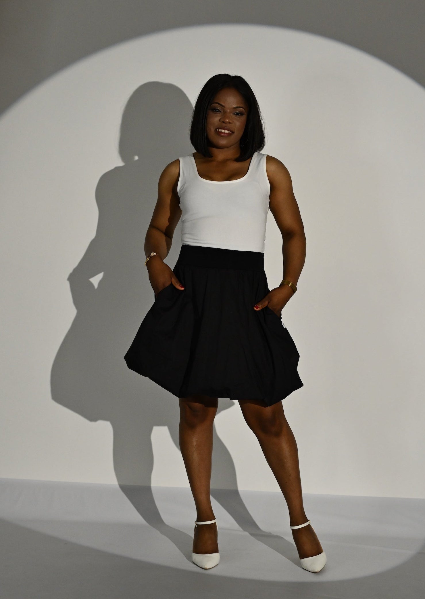 A woman wearing a sleeveless cream top and a black skirt standing in a studio with a spotlight and her shadow cast on the wall.