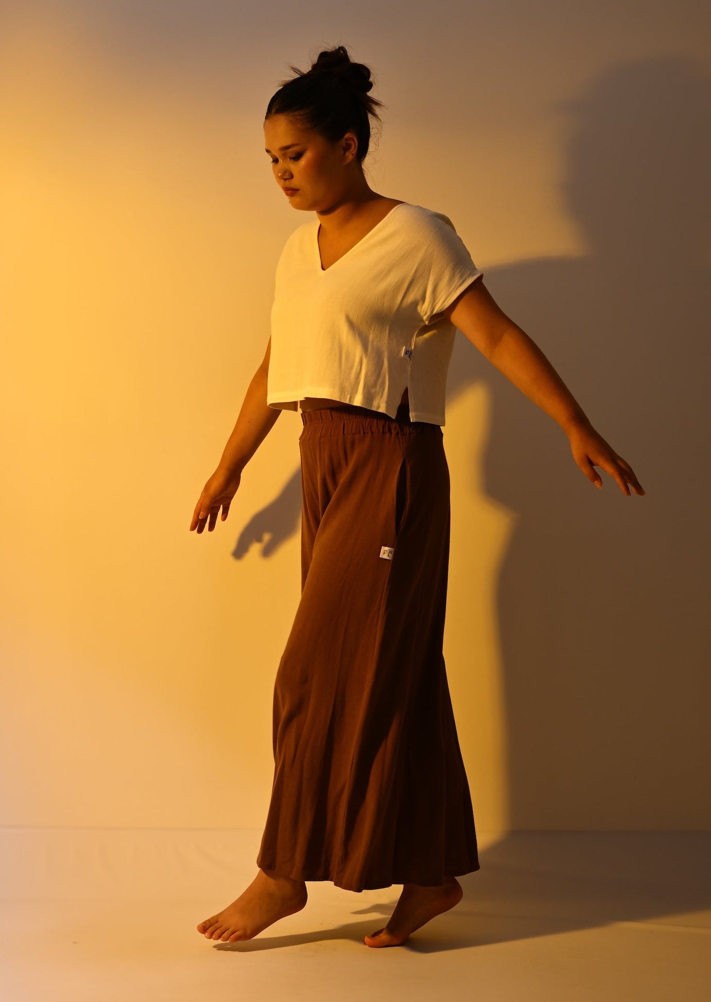A woman wearing a cream cropped short-sleeve top and a brown cropped trousers poses barefoot in a studio with dramatic warm lighting and a strong shadow on the wall.