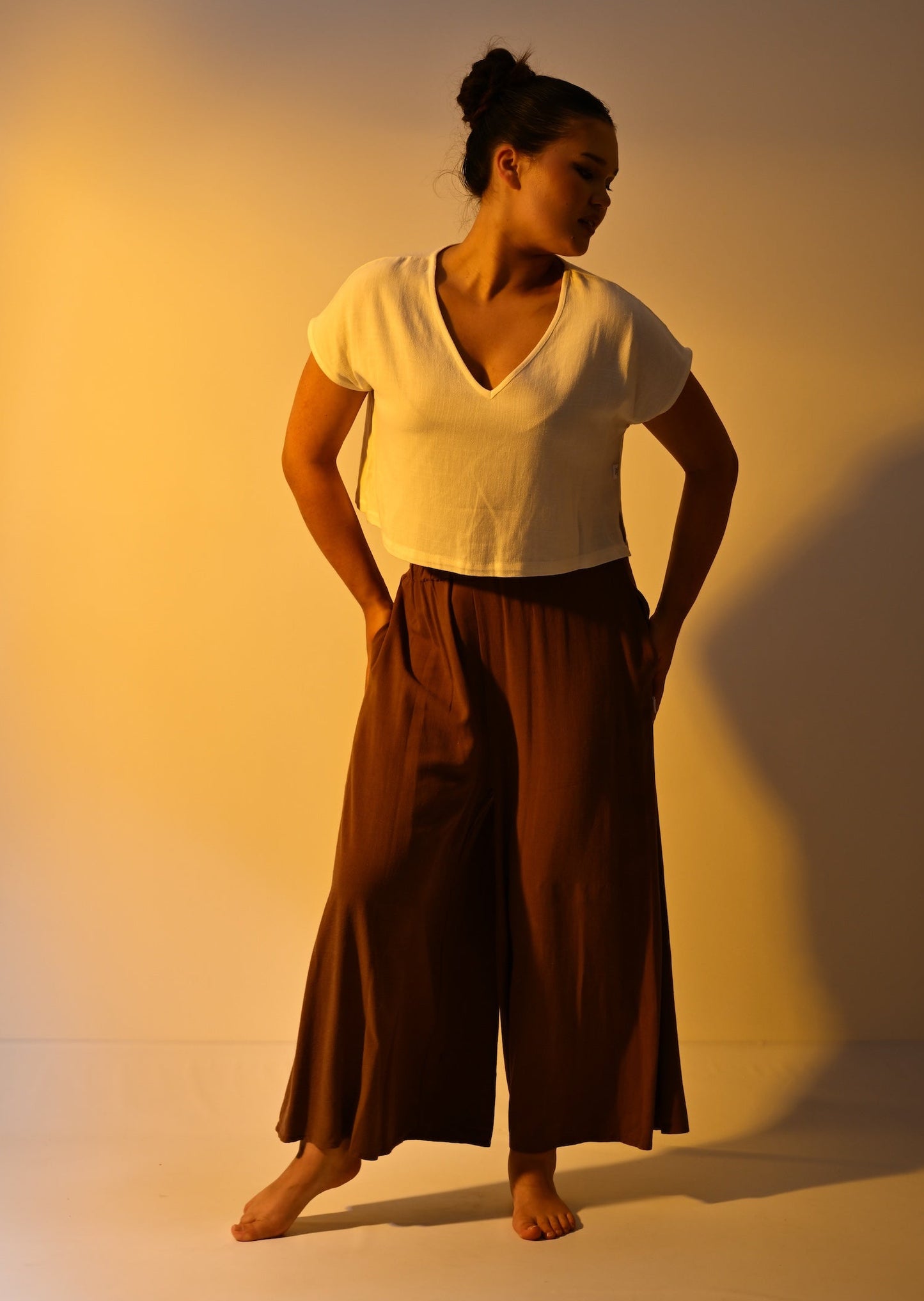 A woman wearing a cream cropped short-sleeve top and a brown cropped trousers poses barefoot in a studio with dramatic warm lighting and a strong shadow on the wall.