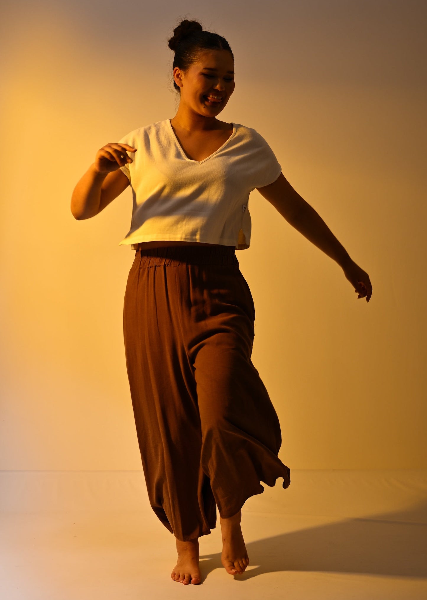 A woman dancing barefoot in a cream short-sleeve crop top and brown wide-leg pants against a warm yellow studio backdrop.