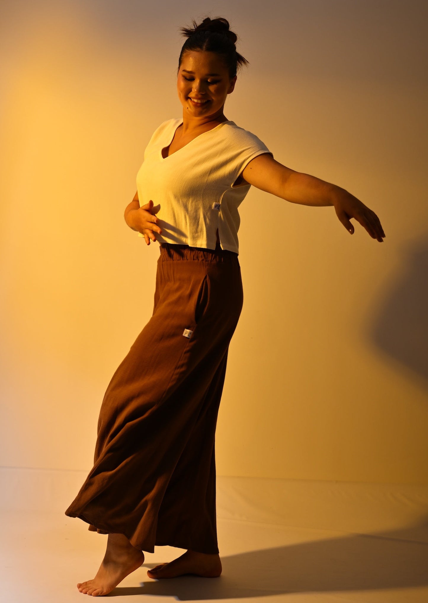 A woman posing in a cream short-sleeve V-neck top and a cropped, flowing brown trousers against a warm, yellow-lit studio backdrop.