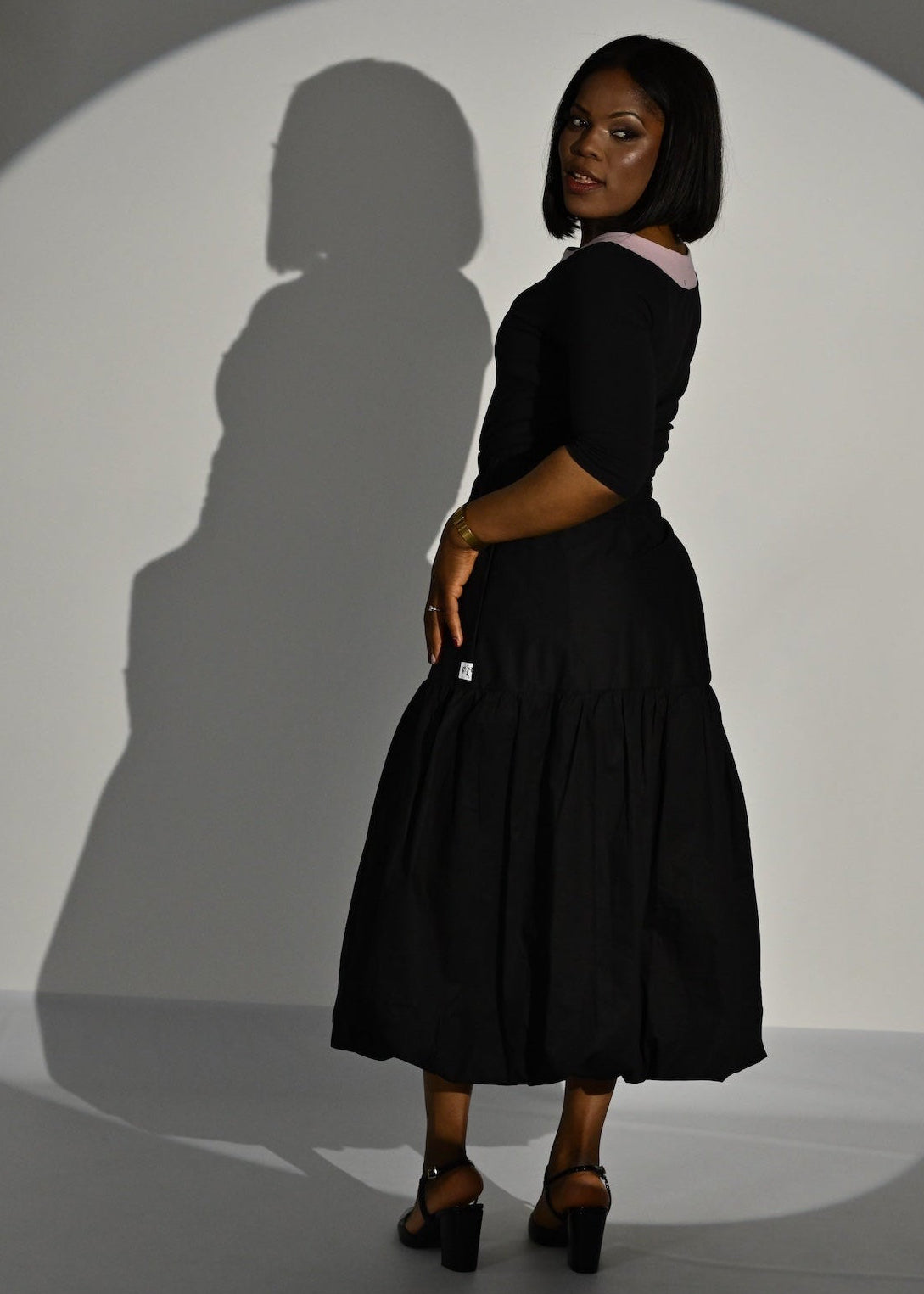 A woman stands in profile wearing a black top with a tiered skirt and black heels, posed in front of a white wall with a large shadow cast behind her.