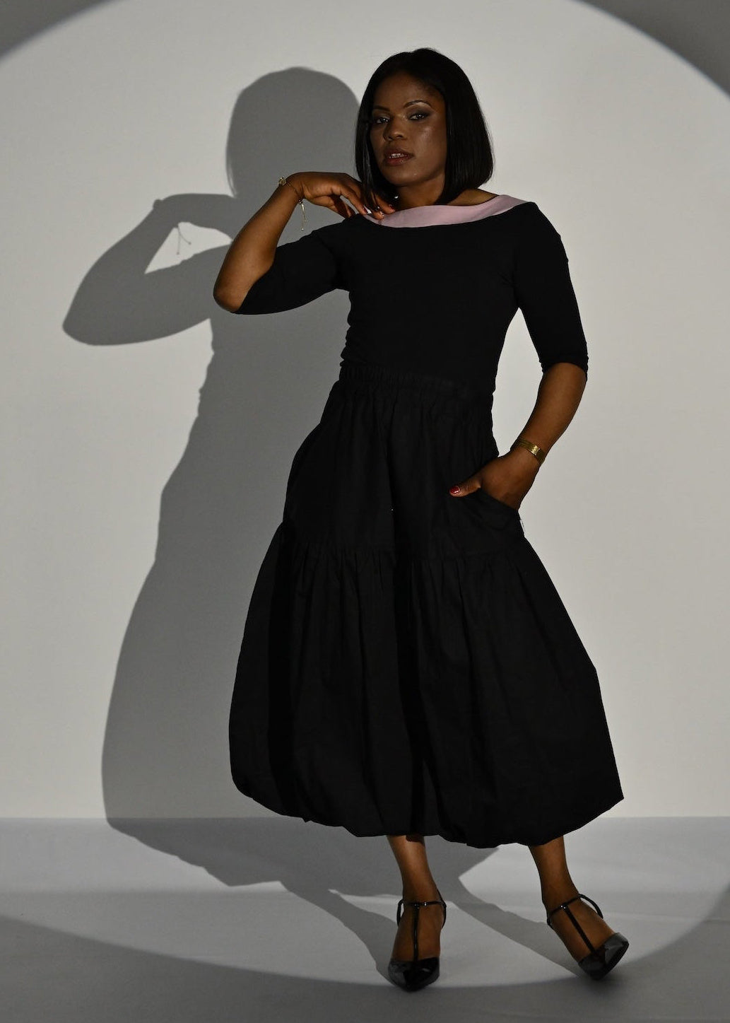 A woman modeling a black midi dress with a wide light pink collar, standing in a studio under a spotlight that casts a long shadow on a white circular backdrop.
