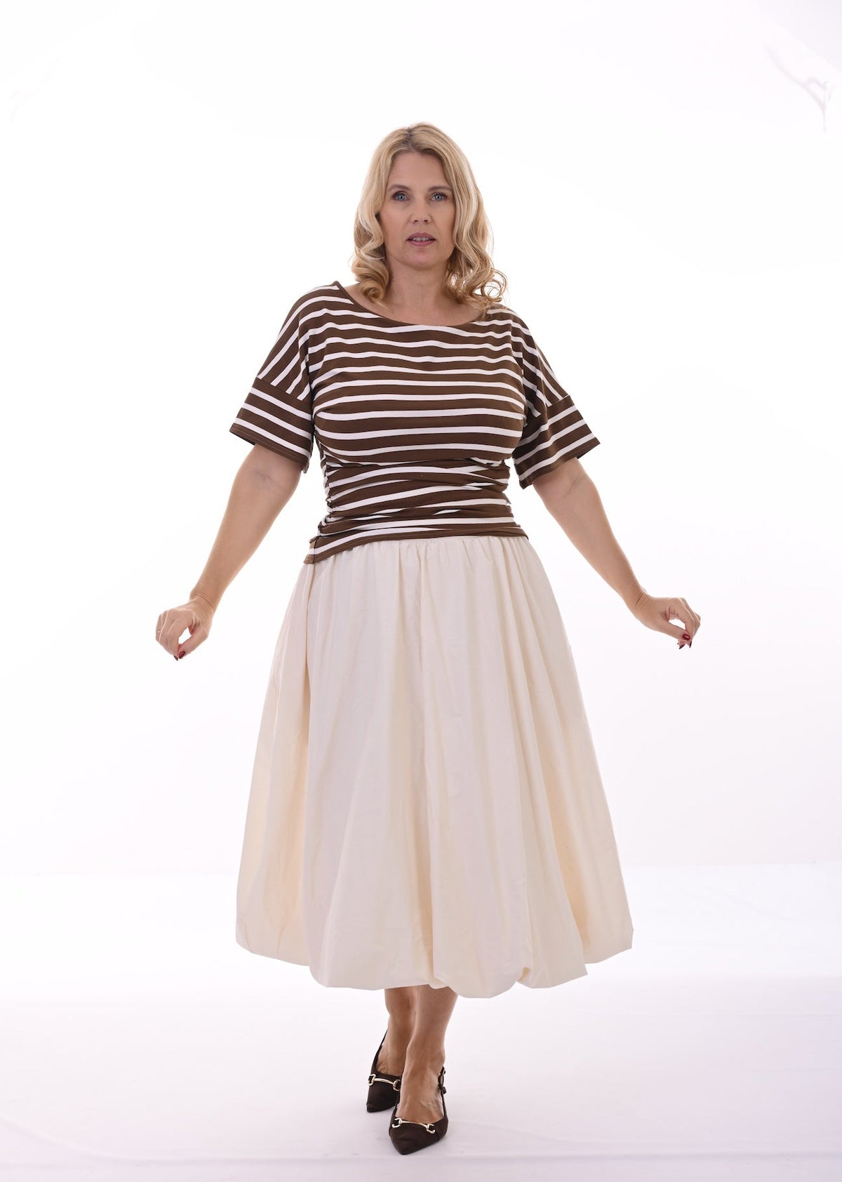 A woman modeling a short-sleeve top with brown and white horizontal stripes, paired with a cream-colored midi skirt and brown heels, against a plain white studio background.