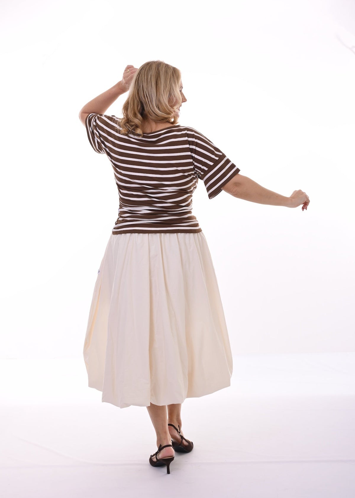A woman is shown from behind wearing a brown and white horizontally striped short-sleeve t-shirt and a pleated cream-colored midi skirt, paired with black strappy heels, against a bright white studio background.