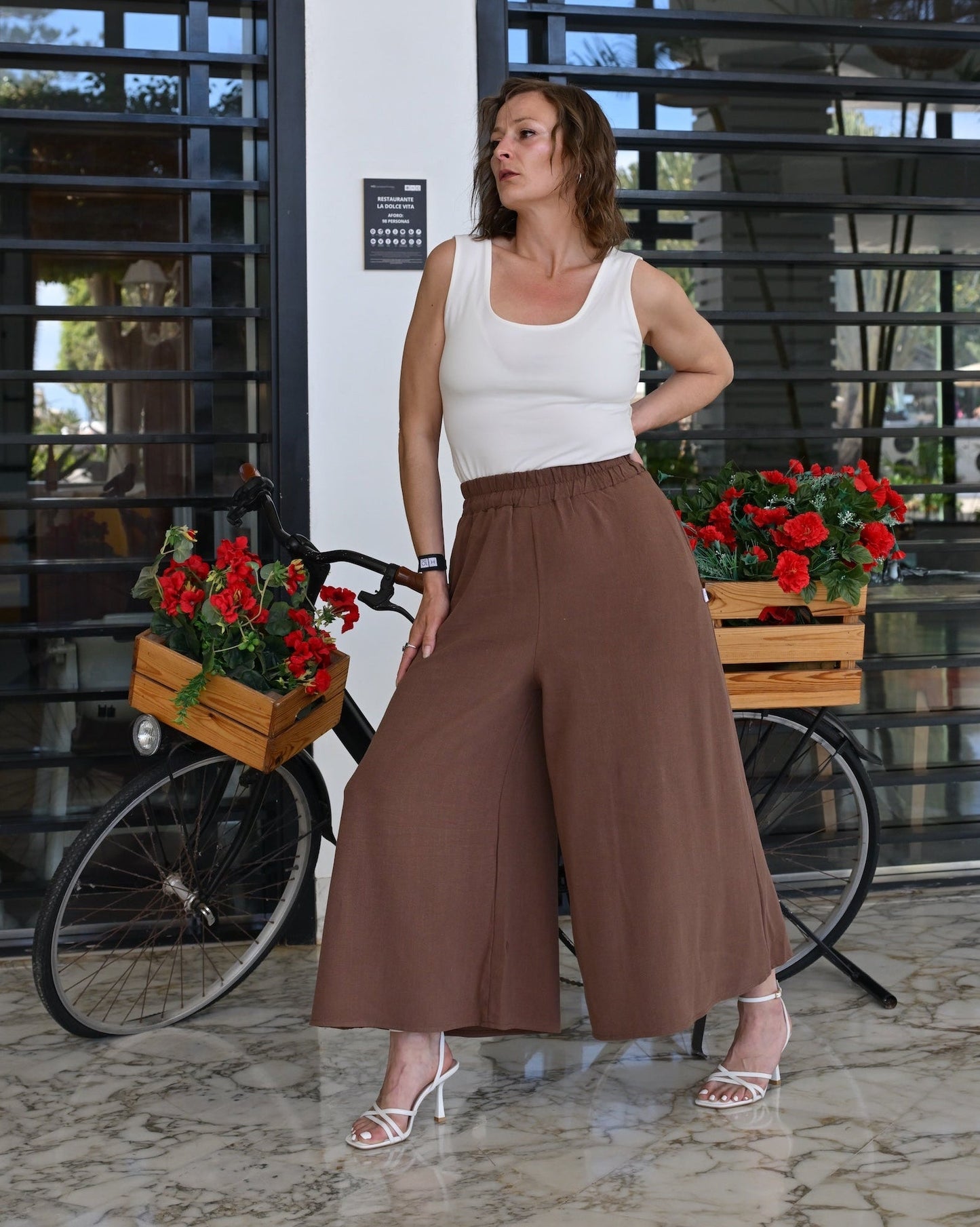 Woman in white top and brown wide-leg pants standing in front of a building with bicycles and flower boxes.