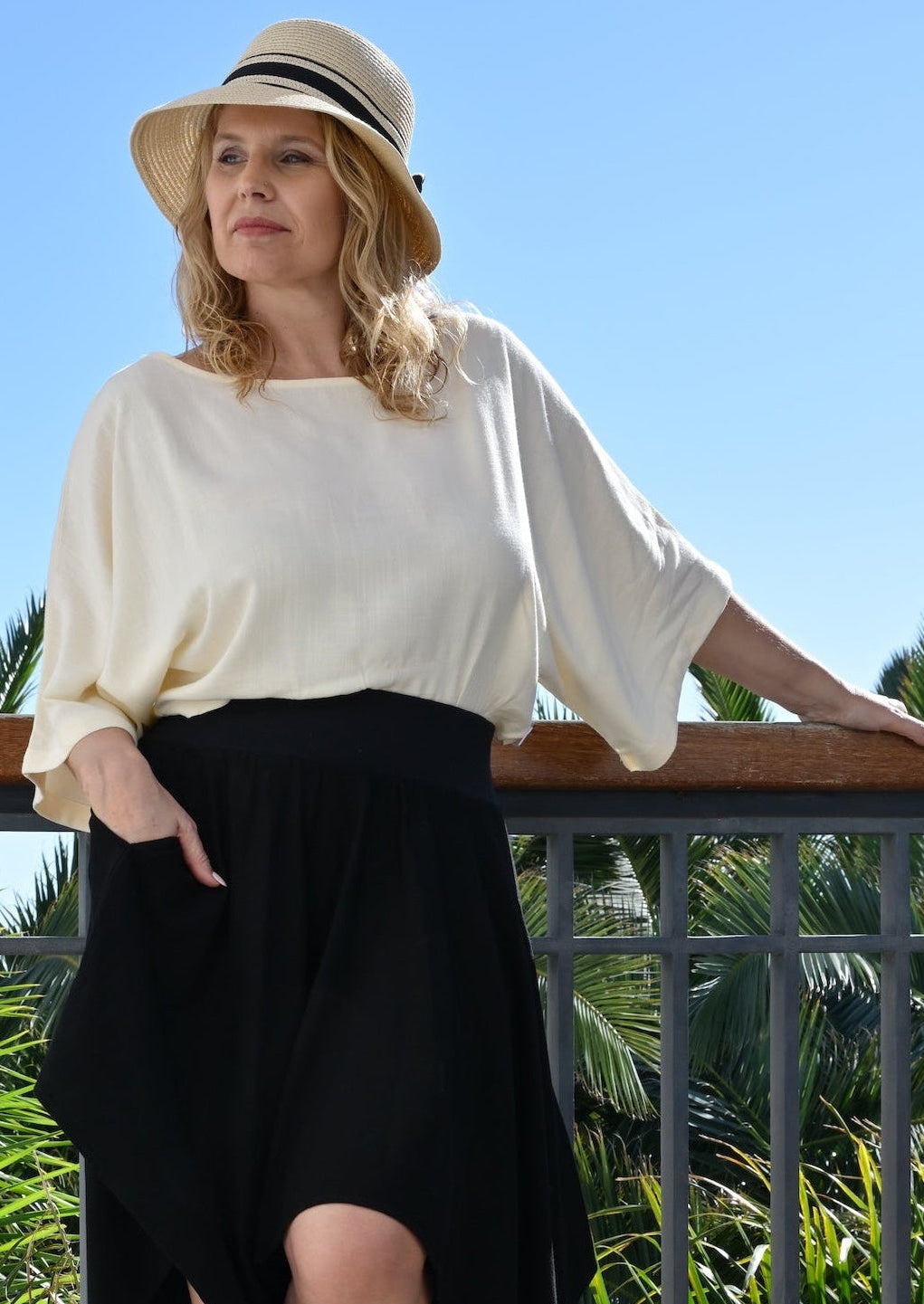 Woman in a white top and black skirt standing on a balcony with palm trees in the background