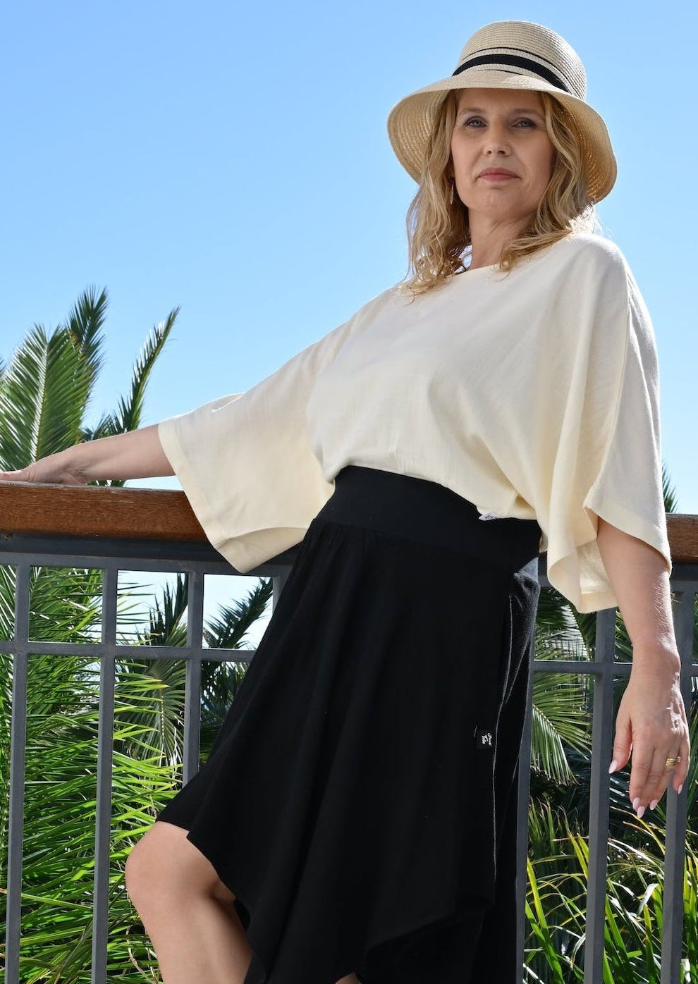 Woman in a white top and black skirt standing on a balcony with palm trees in the background