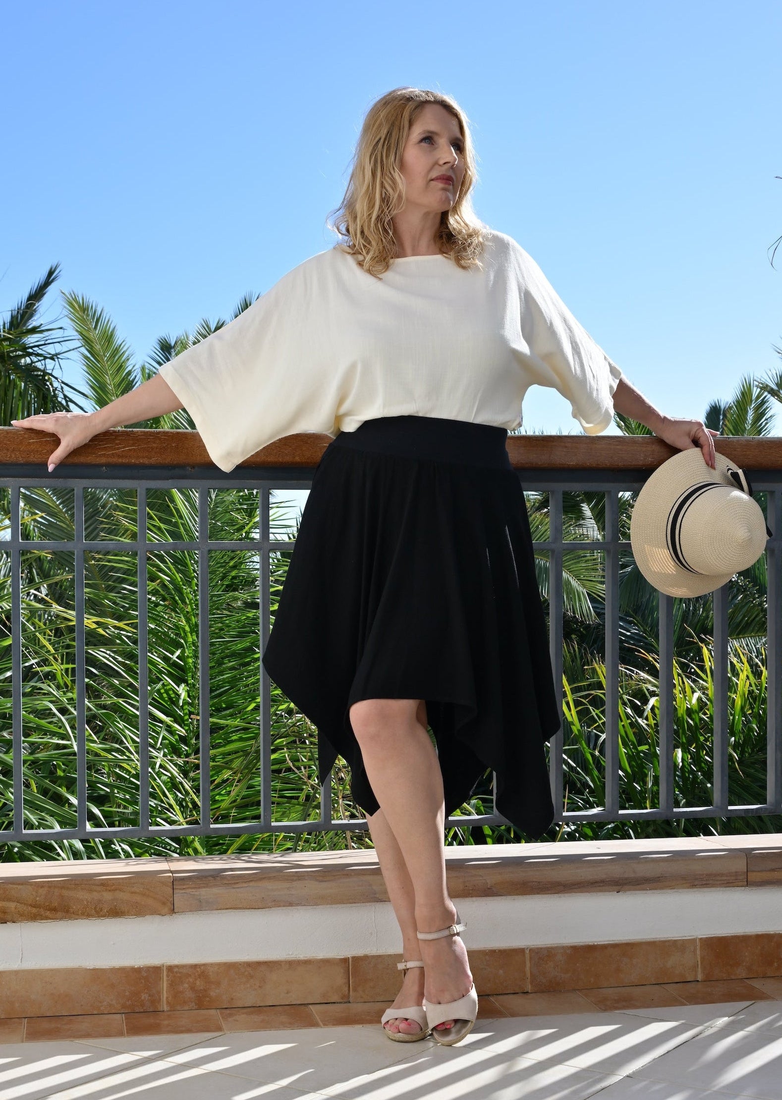 Woman in a white top and black skirt standing on a balcony with palm trees in the background