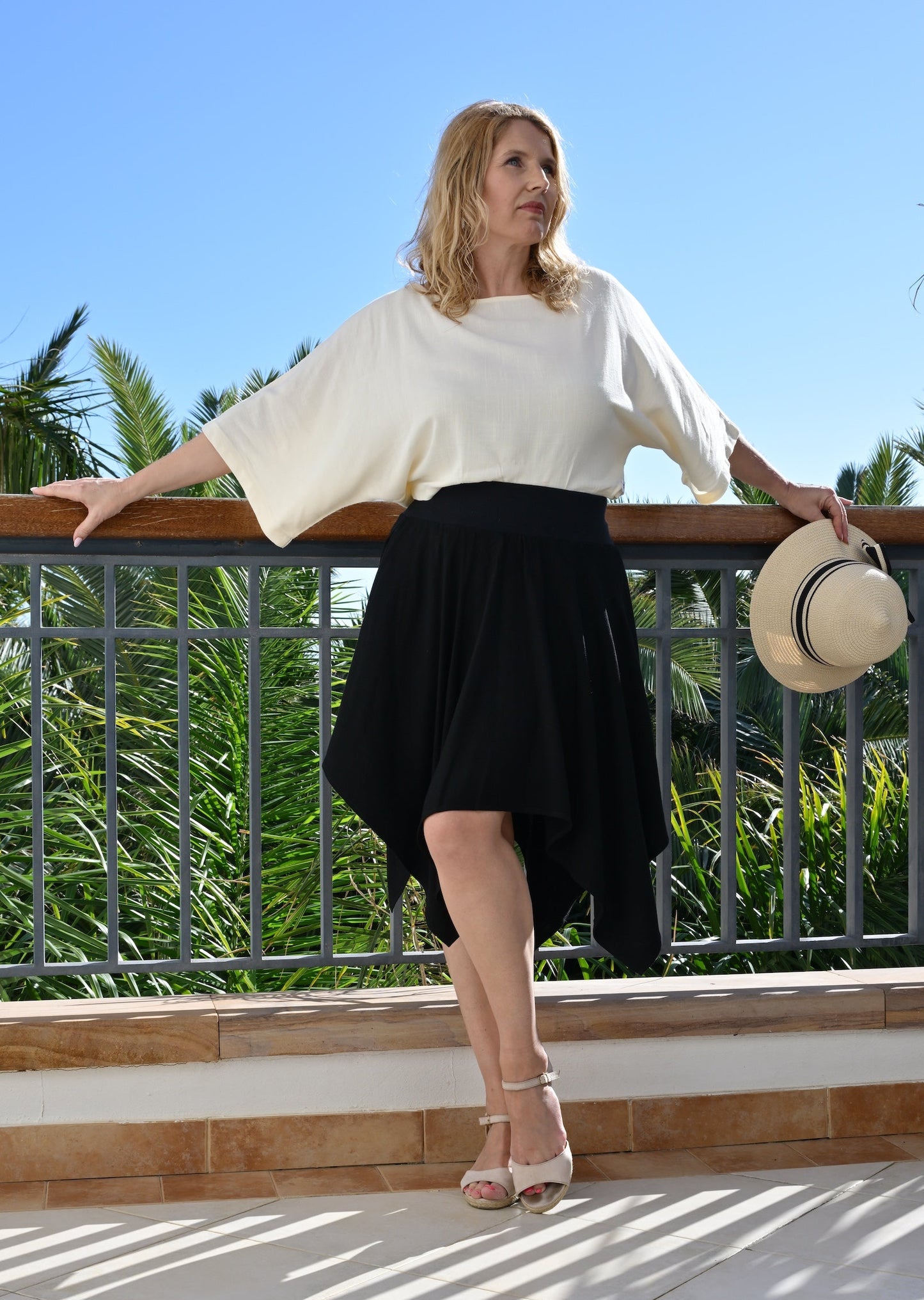 Woman in a white top and black skirt standing on a balcony with palm trees in the background