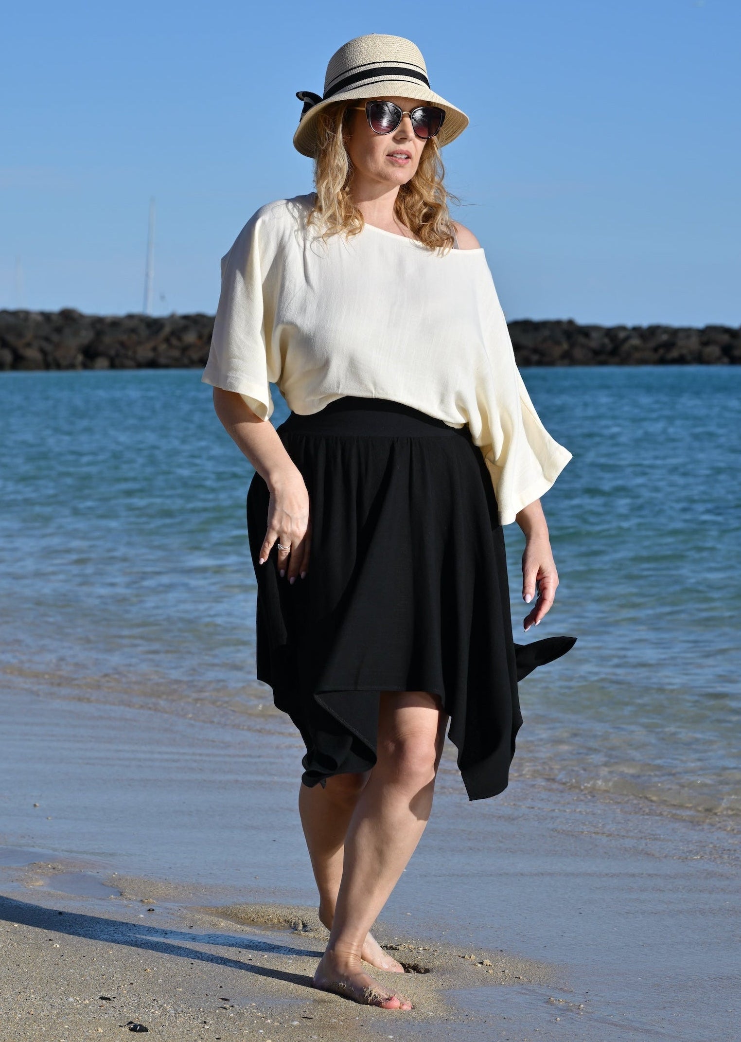 Woman walking on a beach wearing a white top and black skirt with a hat.
