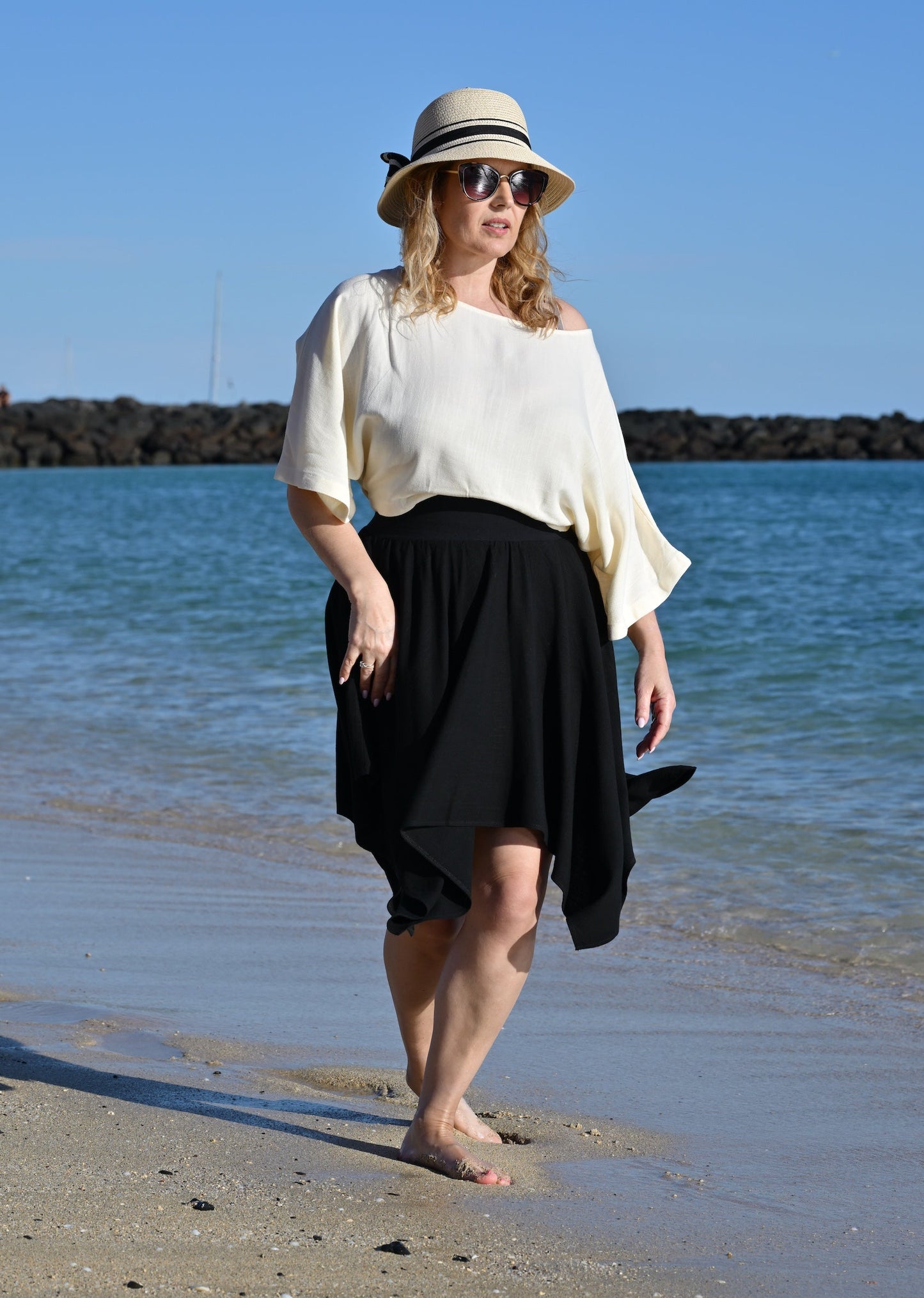 Woman walking on a beach wearing a white top and black skirt with a hat.