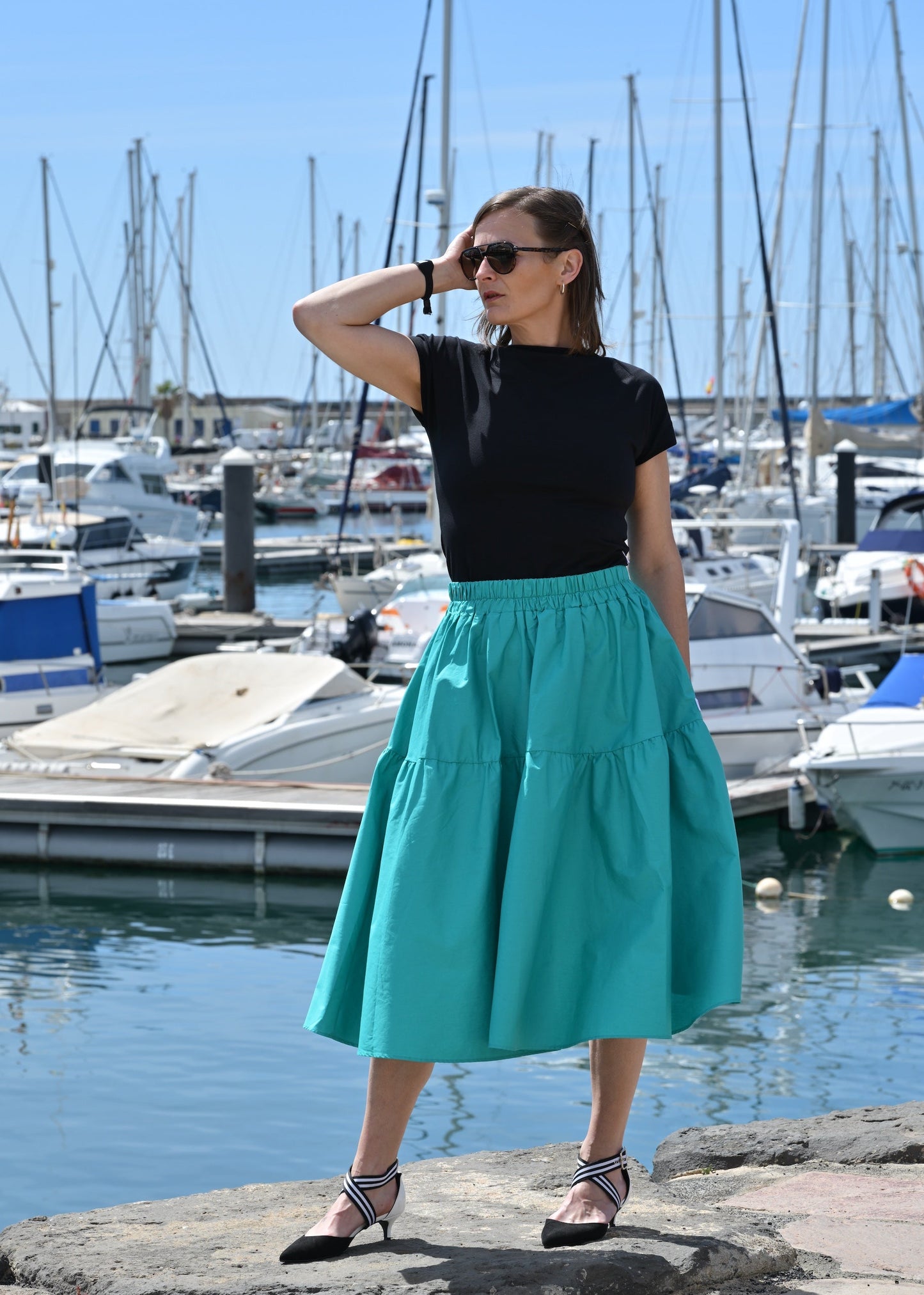 Woman in black top and teal skirt standing by a marina with boats in the background