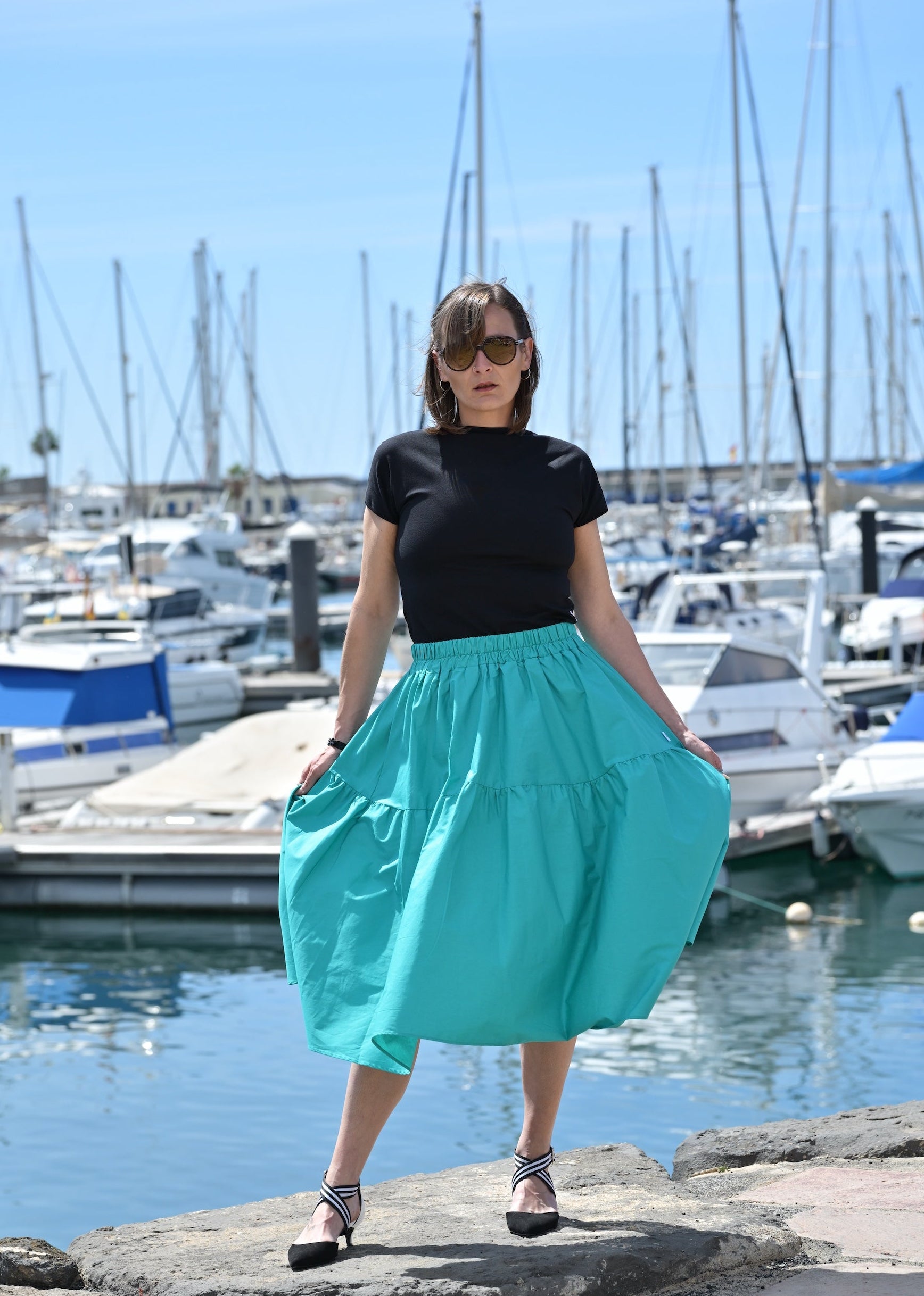 Woman in a black top and teal skirt standing by a marina with boats and clear blue sky.