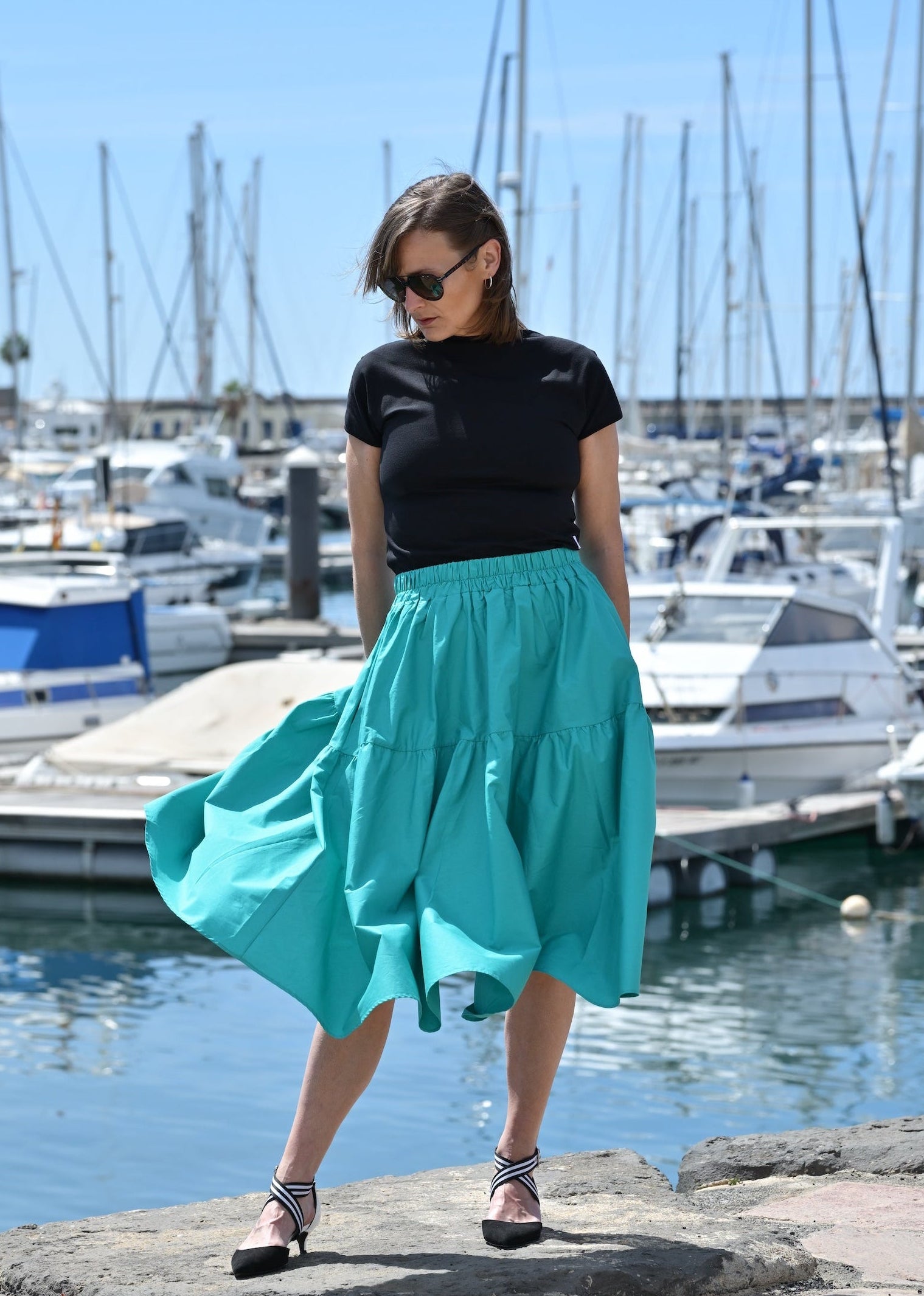 Woman in a black top and teal skirt standing by a marina with boats in the background.
