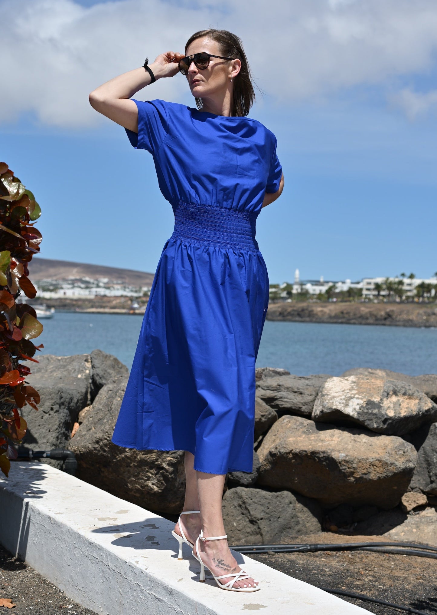 Woman in a blue dress standing by a waterfront with rocks and flowers.