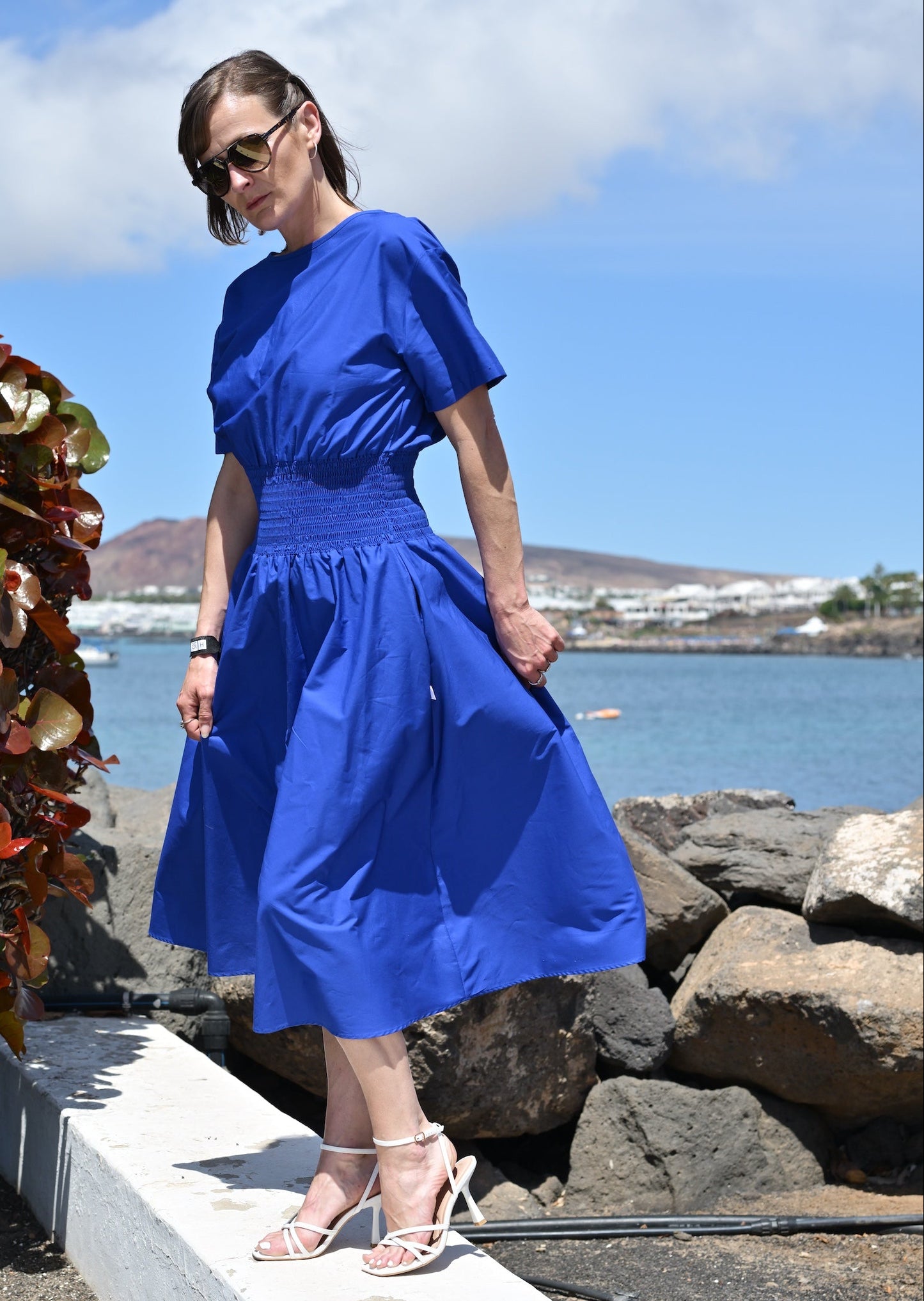 Woman in a blue dress standing by a coastal area with rocks and plants.