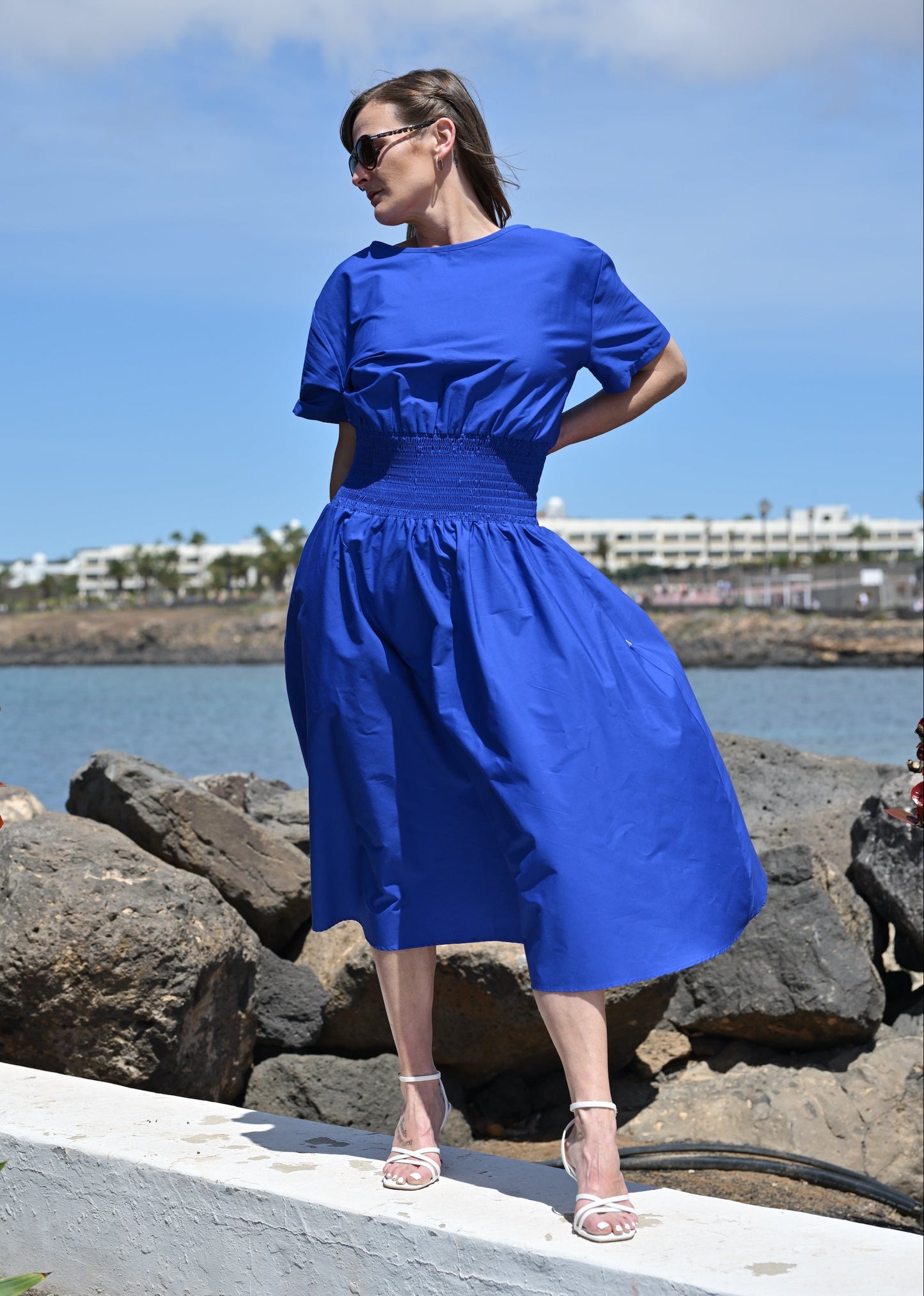 Woman in a blue dress standing on rocks by a waterfront with a clear sky.