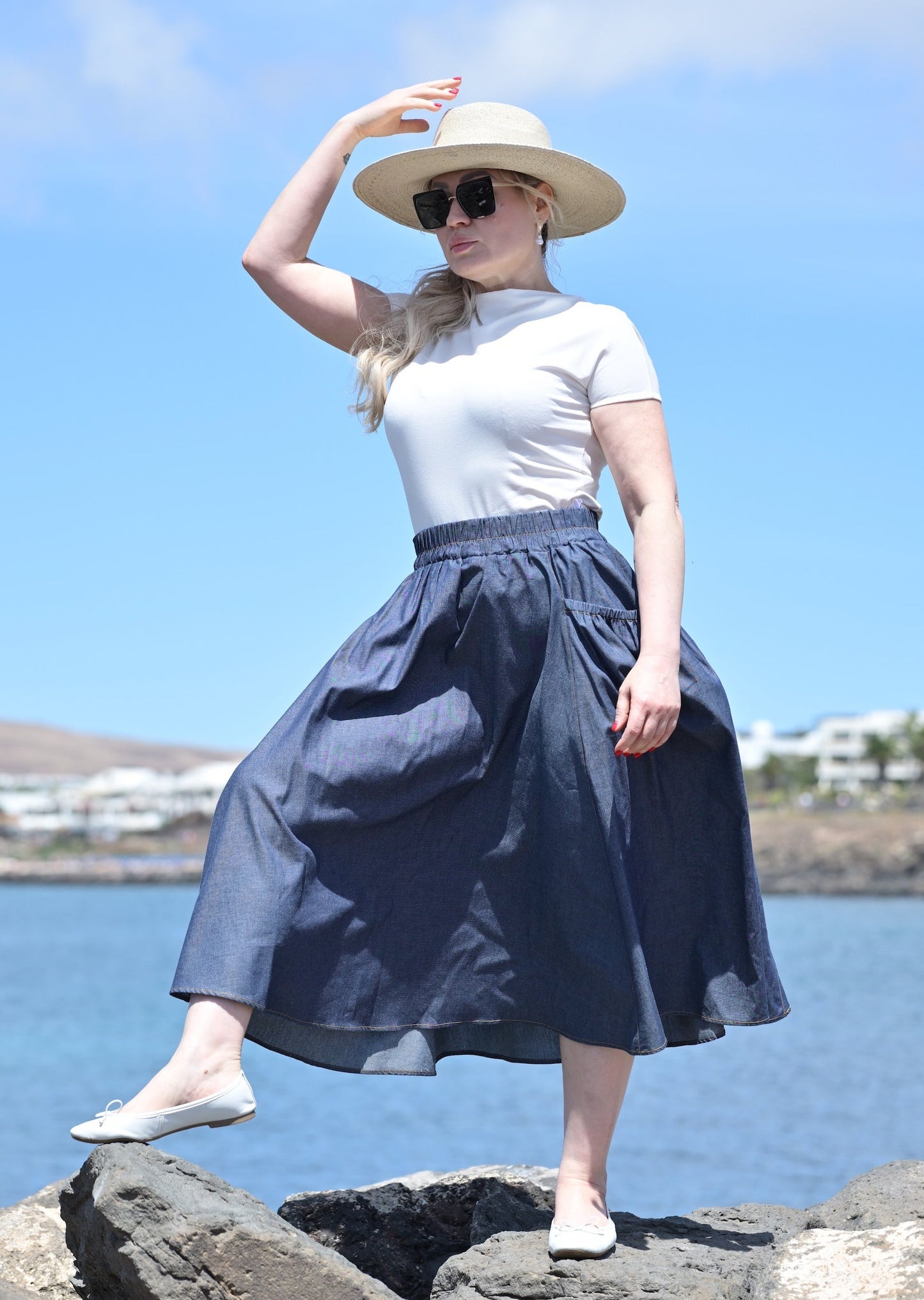 Woman in a white top and blue skirt standing on rocks by a body of water with a blue sky.