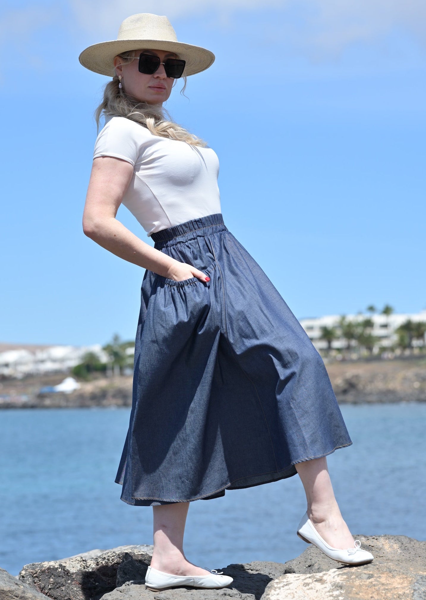 Woman in a white top and blue skirt standing on rocks by a body of water with a blue sky.