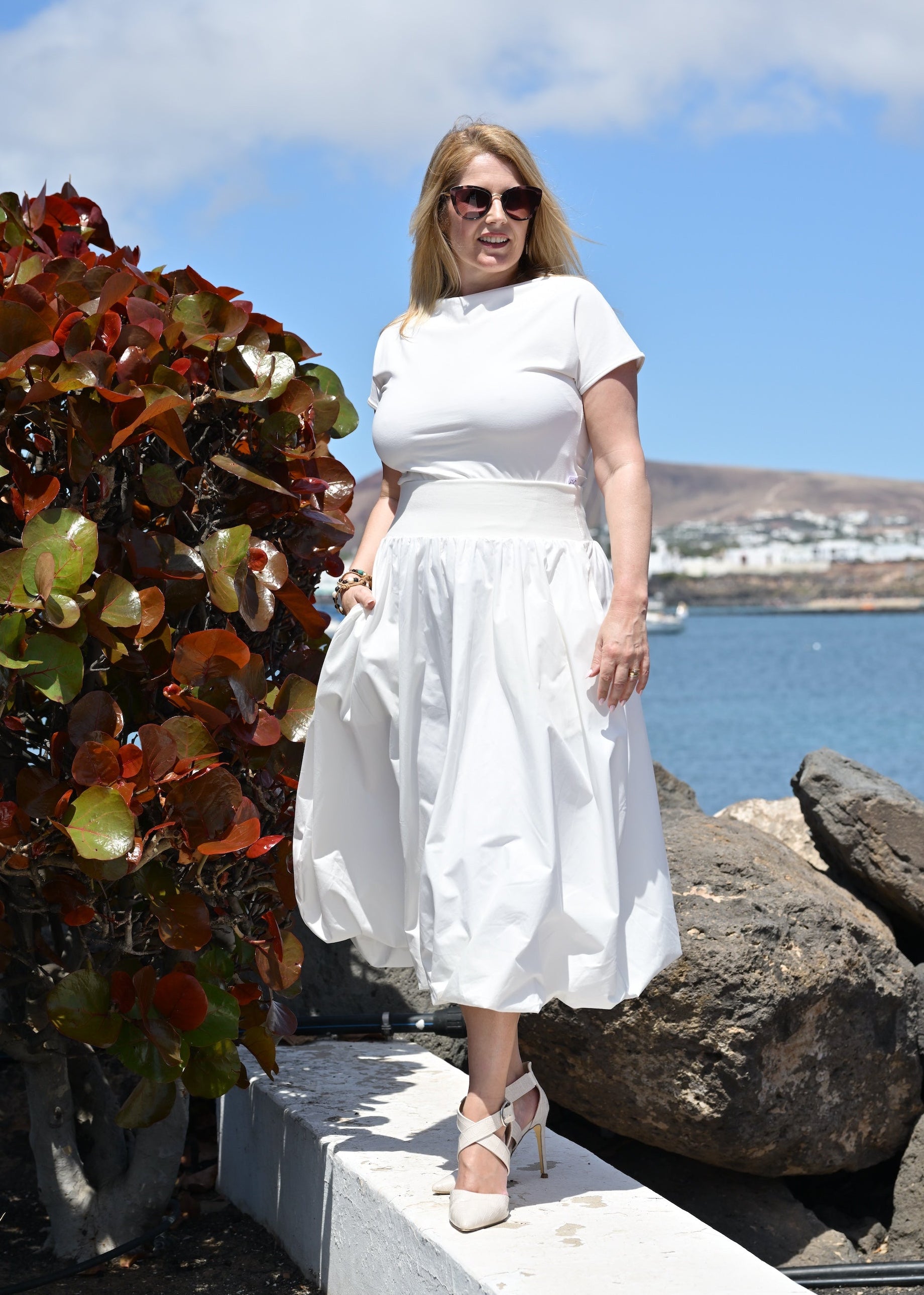 Woman in a white dress standing by a scenic waterfront with colorful foliage.