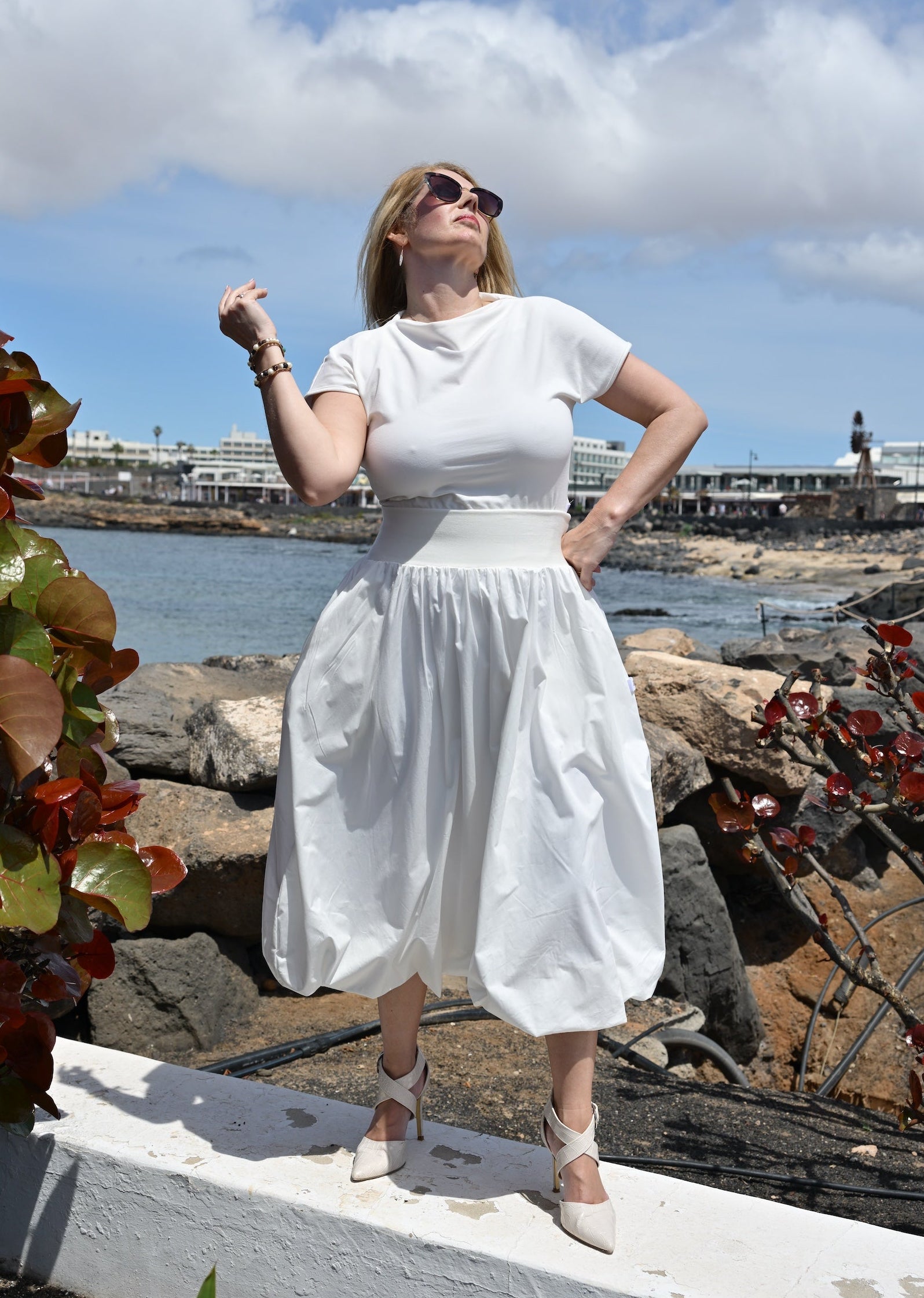 Woman in a white dress standing by a waterfront with rocks and plants.