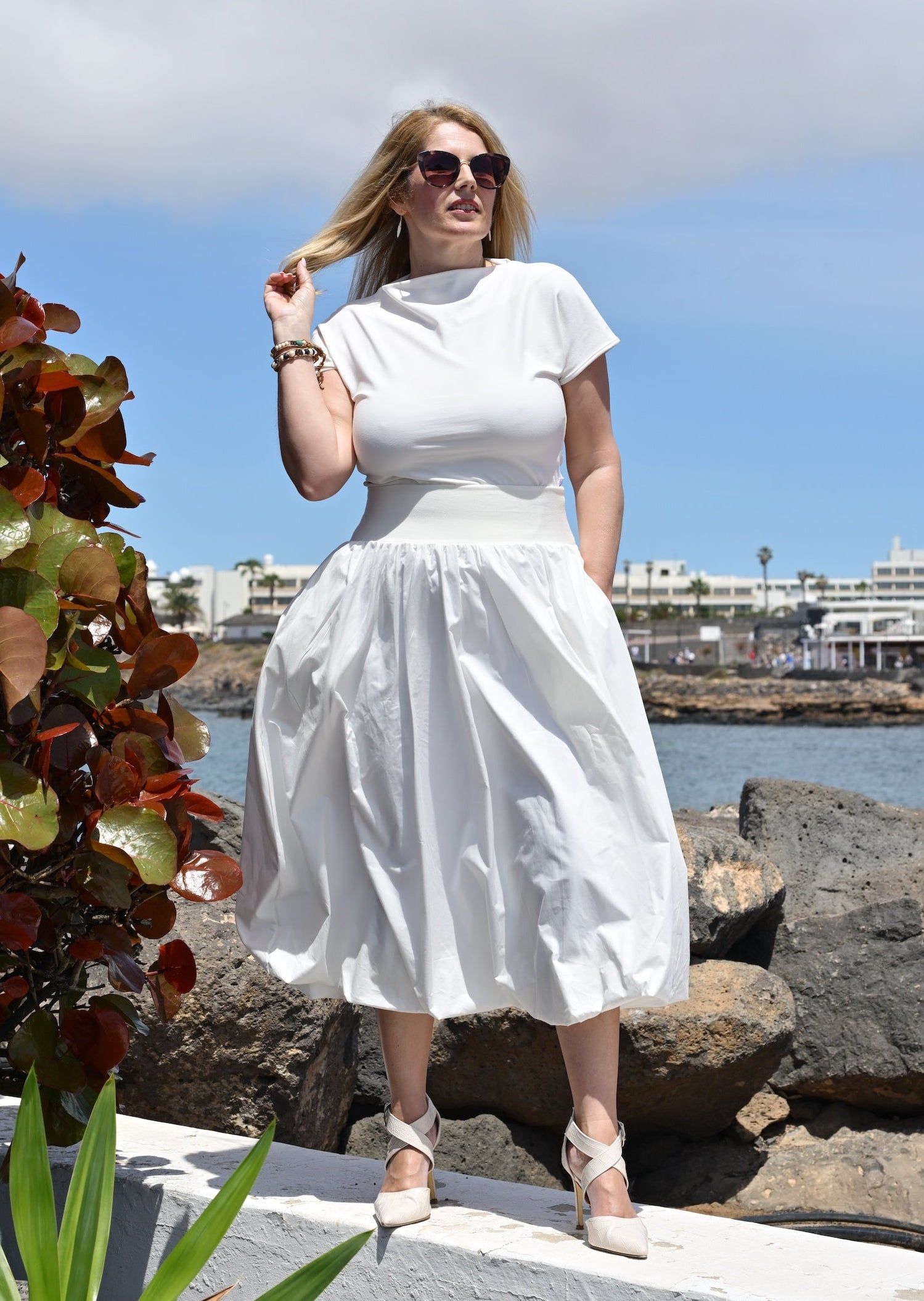 Woman in a white dress standing on rocks by a waterfront with plants and buildings in the background.