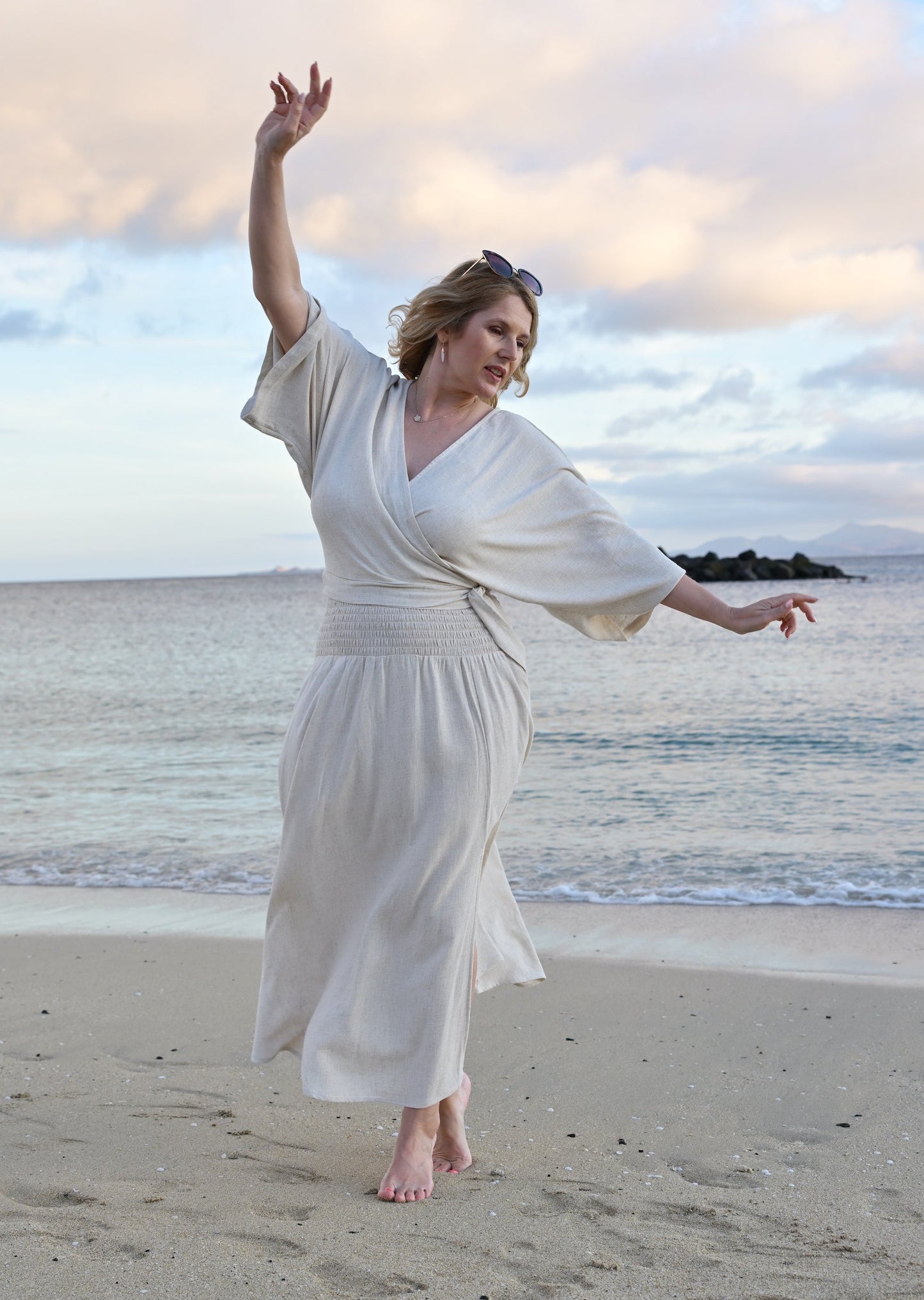 Woman in an oatmeal top and skirt stands standing on a beach with ocean and sky in the background
