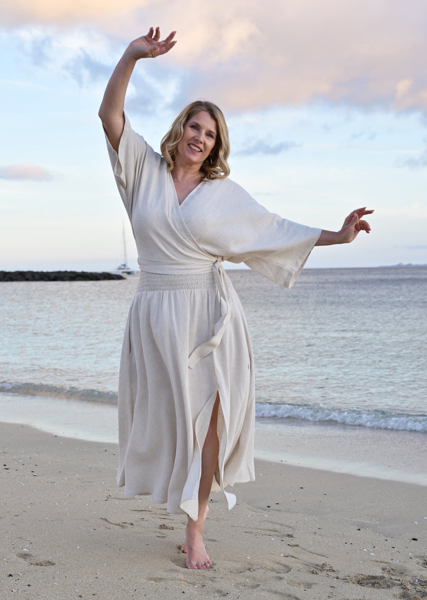 A woman wearing an oatmeal top and skirt stands barefoot on a sandy beach with the ocean and sky in the background.