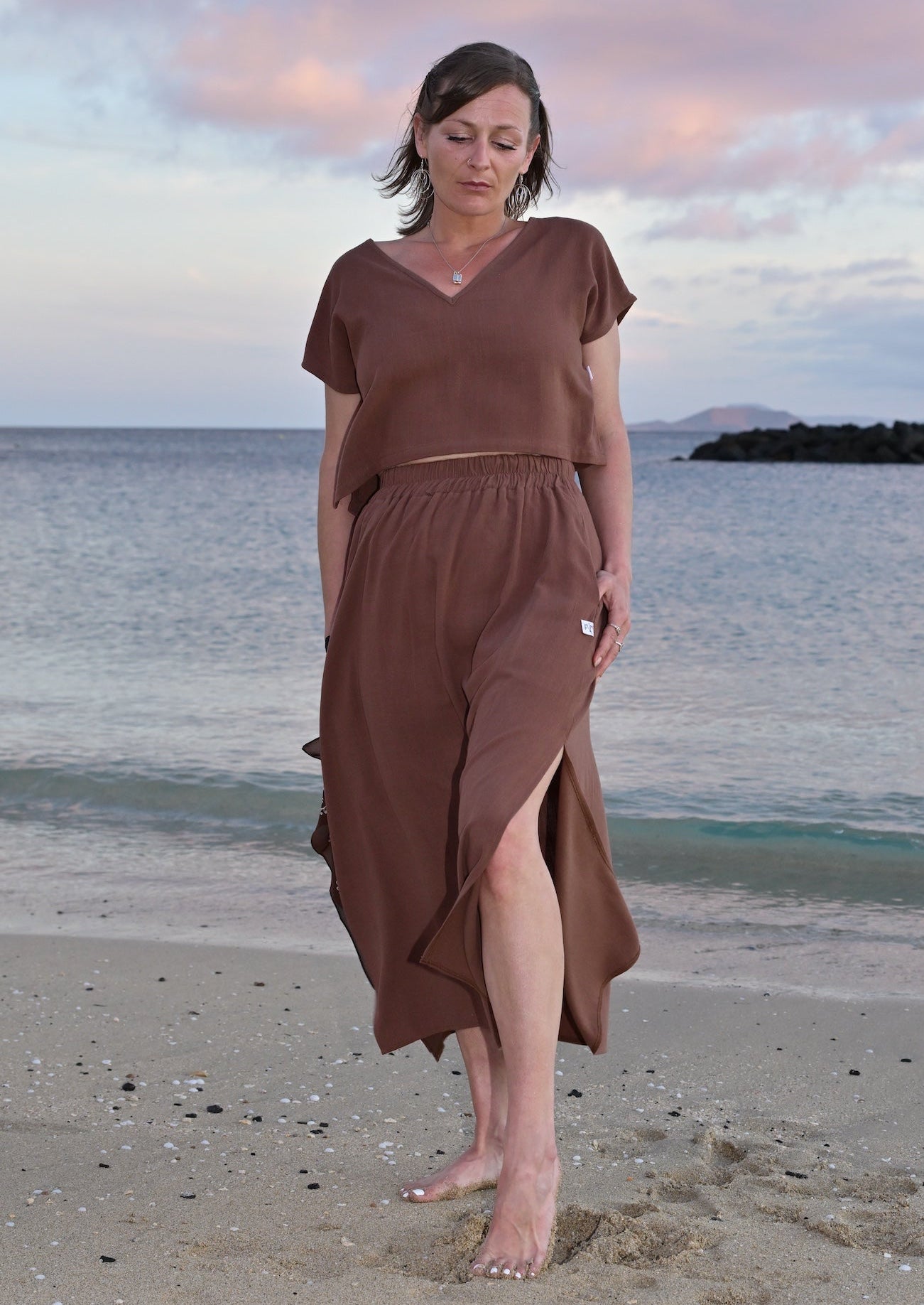 Woman in a brown dress standing on a beach with a sunset sky