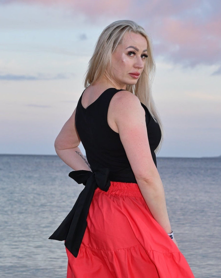 Woman in a black top and red skirt standing on a beach with a sunset sky.
