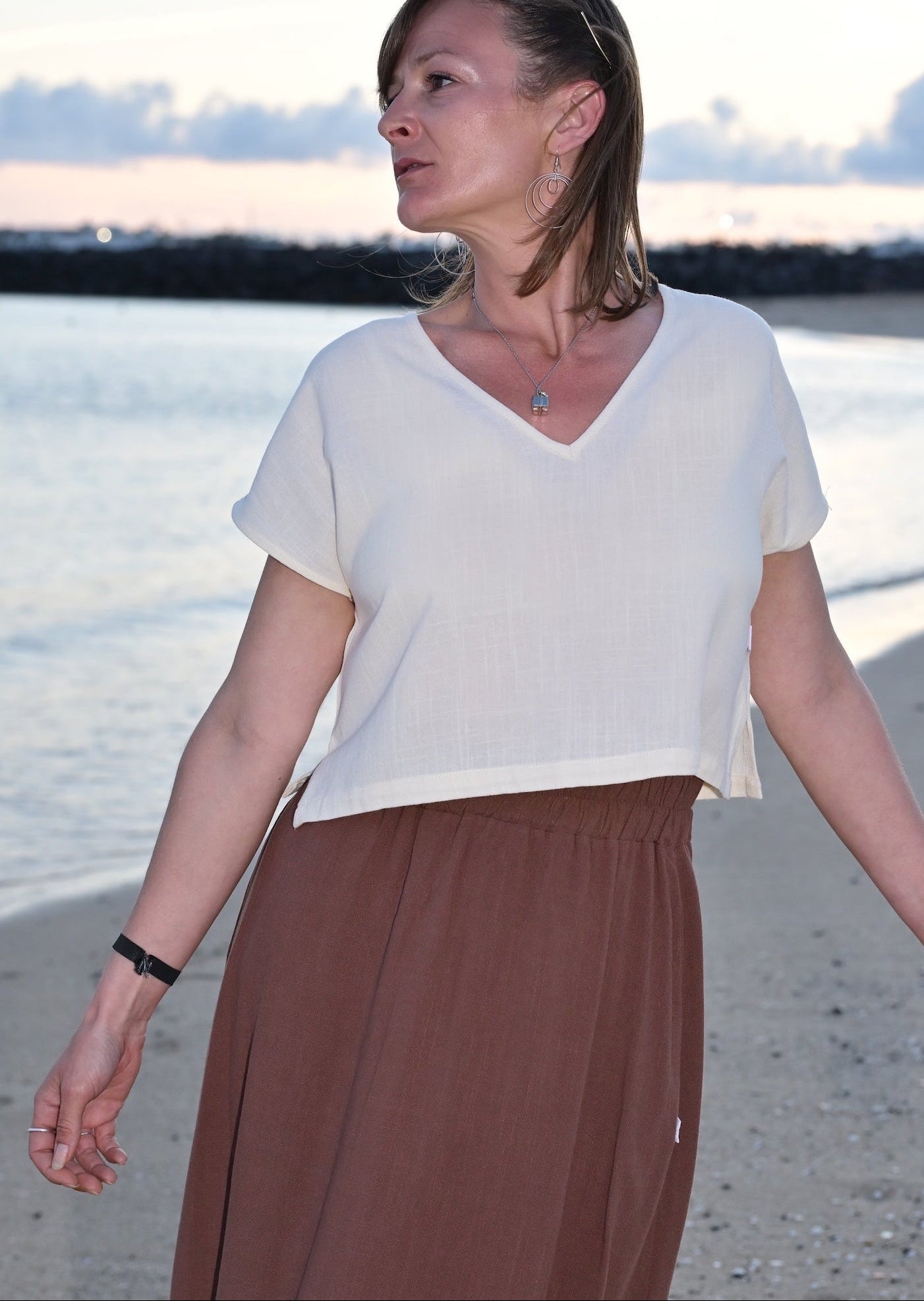 Woman standing on a beach wearing a white top and brown skirt.