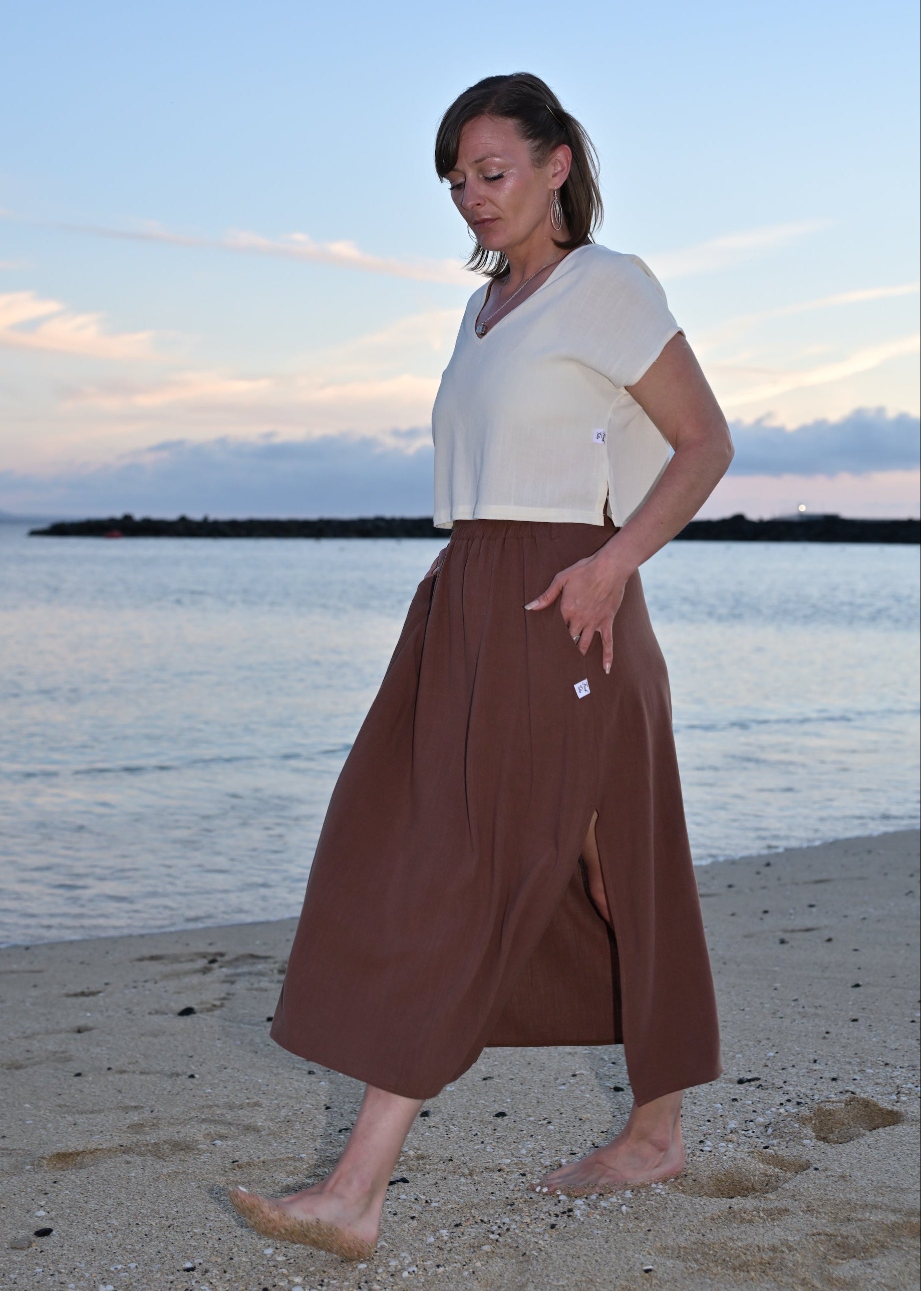 Woman standing on a beach wearing a white top and brown wide-leg pants with a sunset or sunrise in the background.