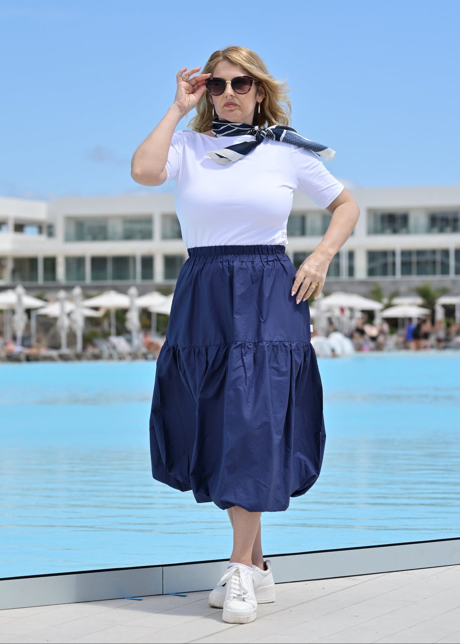 Woman in a white top and navy skirt standing by a pool with a blue sky background