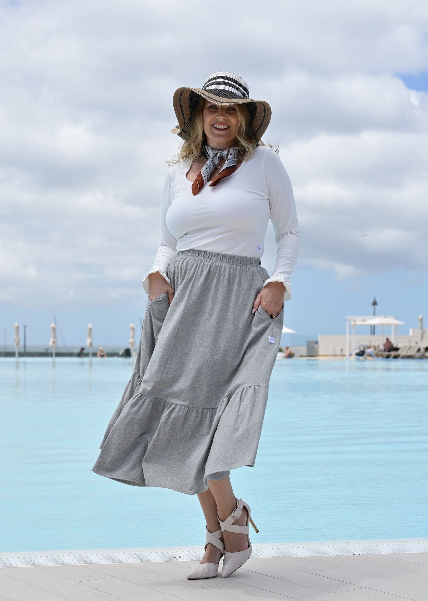 Woman in white top and gray skirt standing by a pool with a cloudy sky.