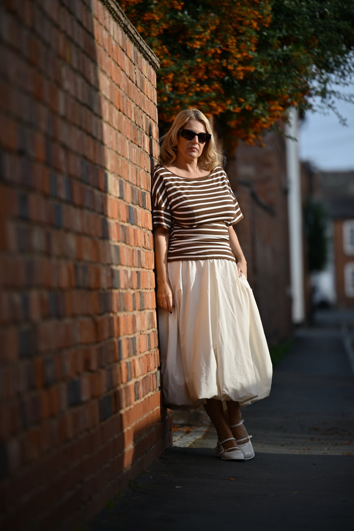 A woman wearing a brown and white horizontally striped short-sleeve top and a voluminous cream skirt, leaning against a red brick wall on a sunny street.