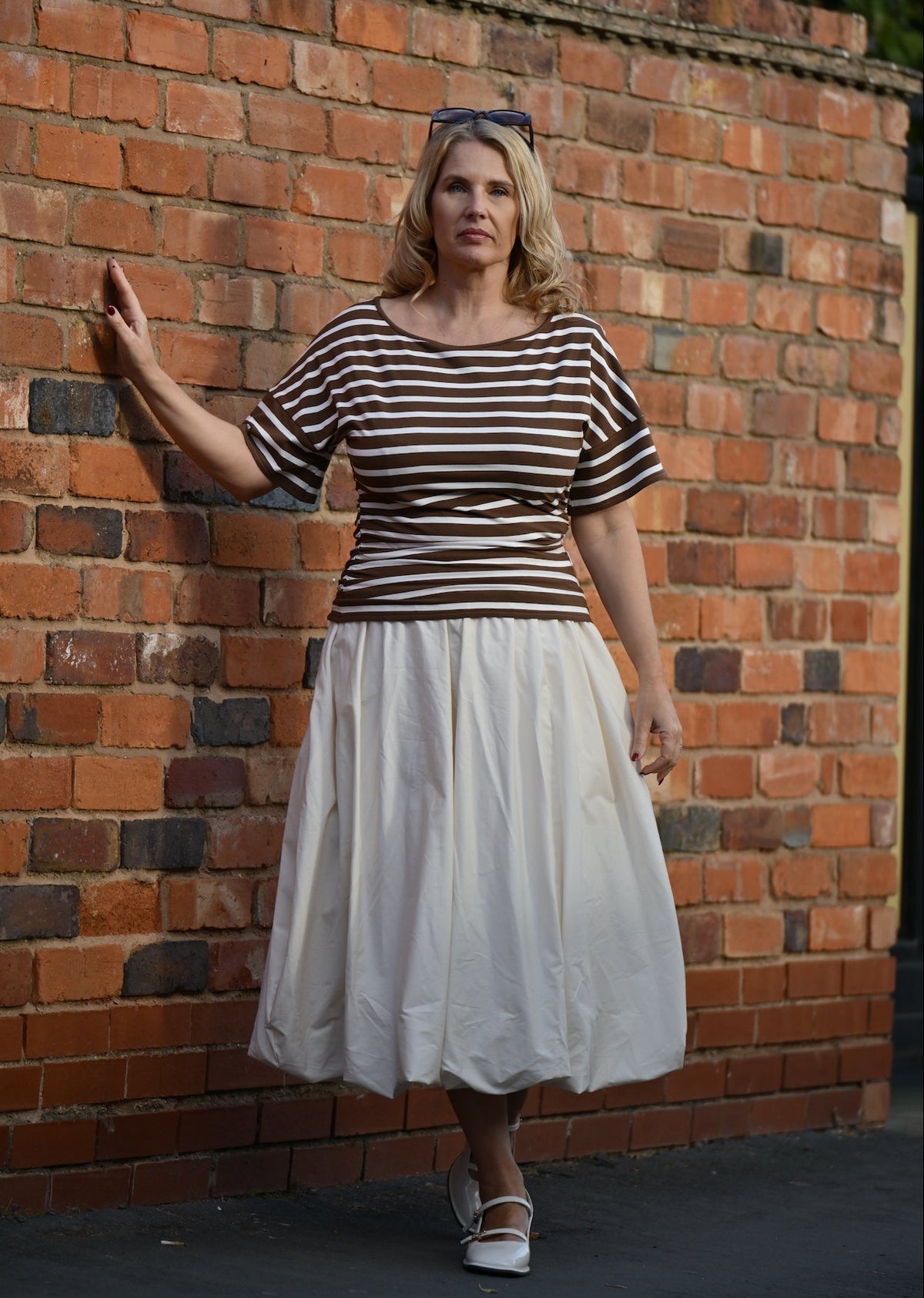 A woman stands against a red brick wall wearing a brown and white horizontally striped short-sleeve top and a light cream pleated midi skirt.