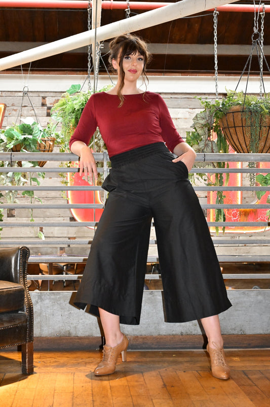 Woman in a red top and black wide-leg pants standing in a room with wooden floors and plants.
