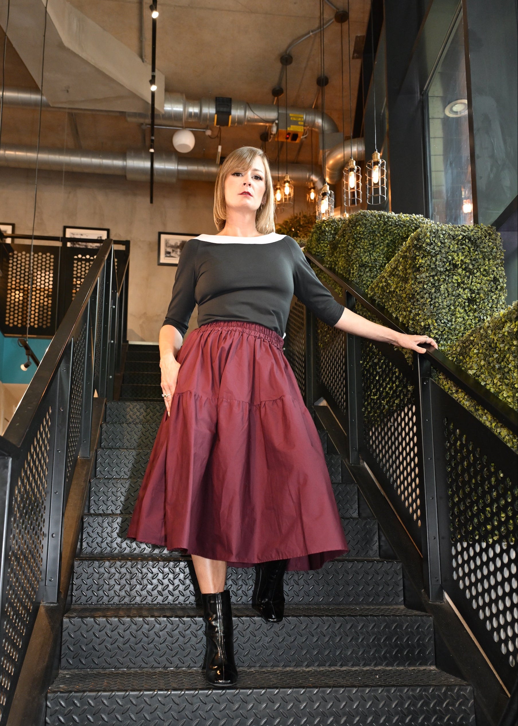 Woman in a maroon skirt and black top standing on a staircase in an indoor setting.