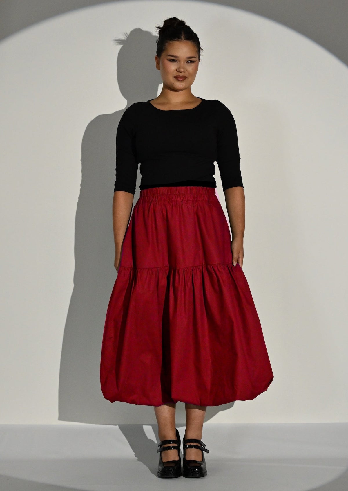 A model stands in a studio spotlight wearing a black three-quarter sleeve top and a voluminous puffball red tiered midi skirt, paired with black Mary Janes.