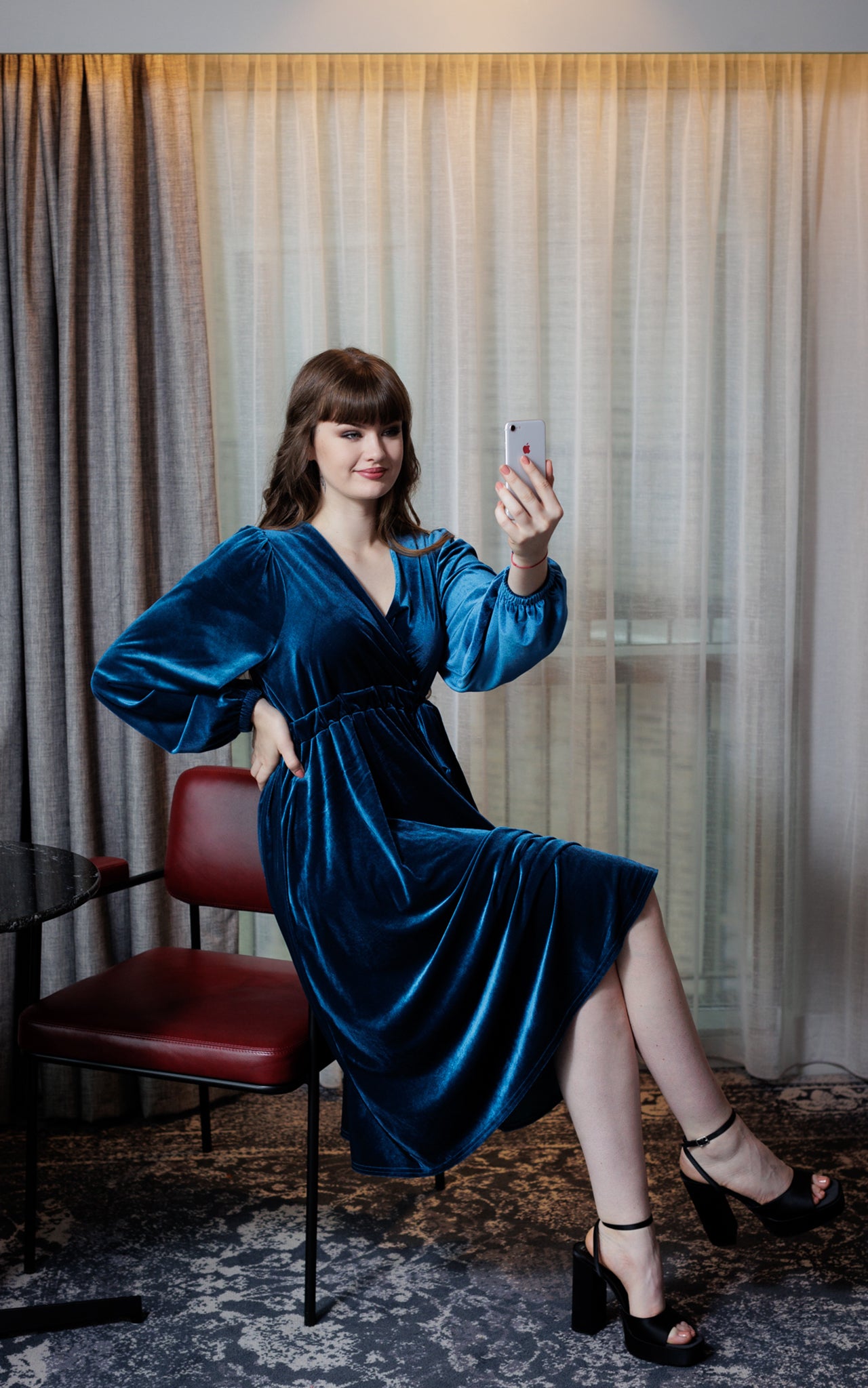 A young ady sits on a chair arm with her right hand on her hip. She holds a phone to her face to take a selfie. She is sitting cross legged and wears black sandals. Her dress is teal blue velvet with a v neck and gathered waist. The skirt is full and flowing. The background is sheer natural coloured curtains.