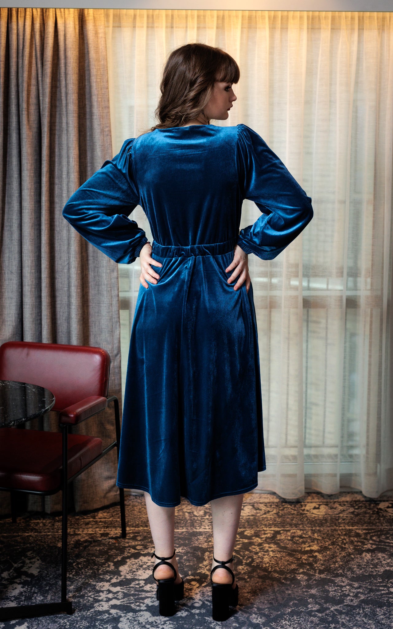 This image is a back view of the model wearing the teal velvet dress. The lighting highlights the gathers on the dress and the shoulder-line. The model has brown hair and is wearing black heels. The background is a cream and grey curtain and the model is standing on a cream and grey patterned rug. There is also a maroon coloured chair on the left side of the image. 