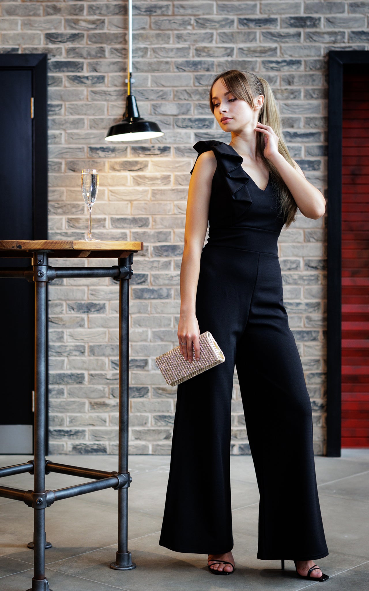 This is a young lady wearing a black V neck jumpsuit with wide legs and a bow on the shoulder. She is wearing black strappy high heels and holding a sparkly clutch bag. She is silhouetted against a brick wall, with her right hand on her neck and looking down her left. 