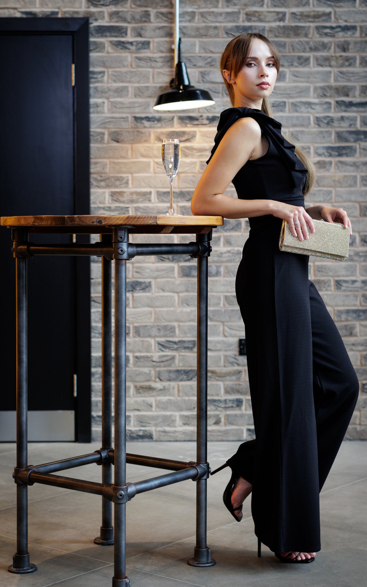 This is a young lady wearing a black wide leg jumpsuit with a bow on the shoulder. She is wearing black strappy heels and holding a sparkly clutch bag. She is silhouetted against a brick wall, and leaning on a tall table with a glass of champagne.