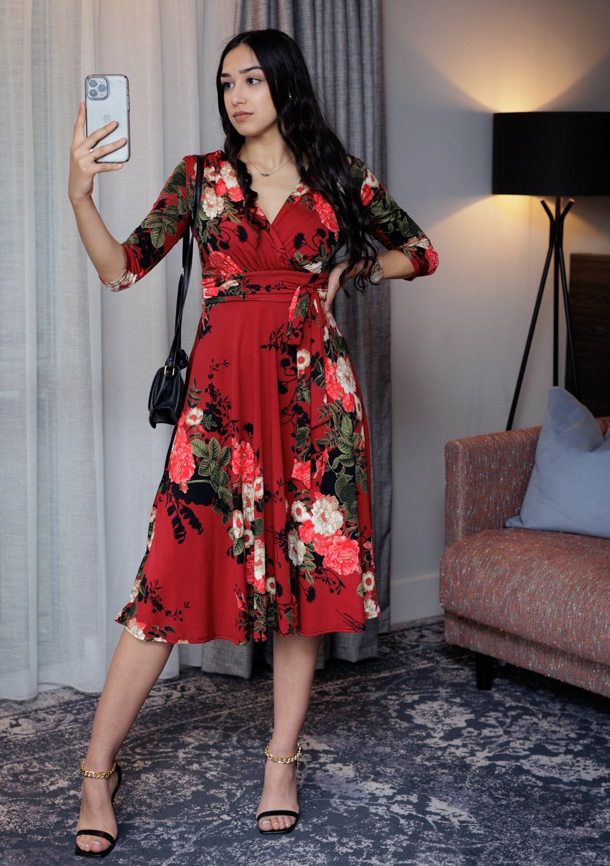 This is a young lady wearing a Mock wrap midi dress in a Red floral design, with 3/4 length sleeves, a gathered shoulder and under bust area, with a tie, secured in a bow. It has a V neck front. The dress has a midi length full skirt in a retro style. The model is wearing black strappy heels, with a small black shoulder bag. She has long dark hair and is taking a picture, she is silhouetted against a modern furnished room.