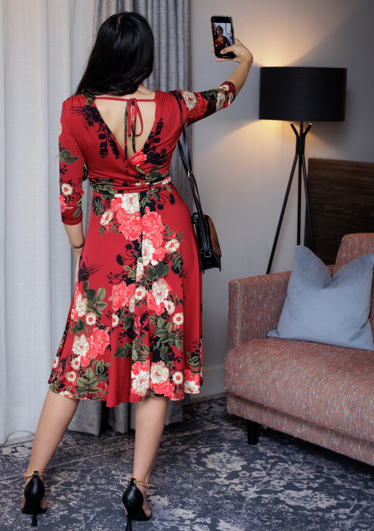 This is a young lady wearing a Mock wrap midi dress in a Red floral design, with 3/4 length sleeves, a gathered shoulder and under bust area, with a tie, secured in a bow. It has a V shaped back with a bow securing it across the top. The dress has a midi length full skirt in a retro style. The model is wearing black strappy heels, with a small black shoulder bag. She has long dark hair and is taking a picture, she is silhouetted against a modern furnished room.